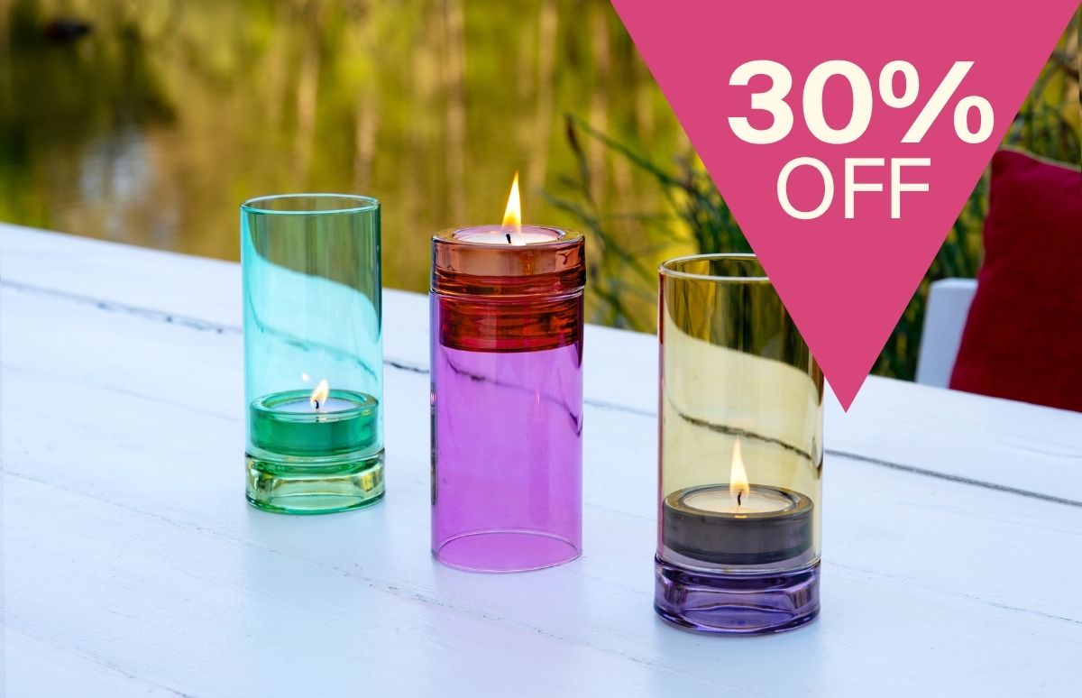 3 farbige Glas-Kerzenhalter mit brennenden Teelichtern auf weißem Holztisch draußen (teal, violett/orange, gold/lila), Pflanzen & Wasser im Hintergrund. Pink: „30% OFF“ oben rechts.