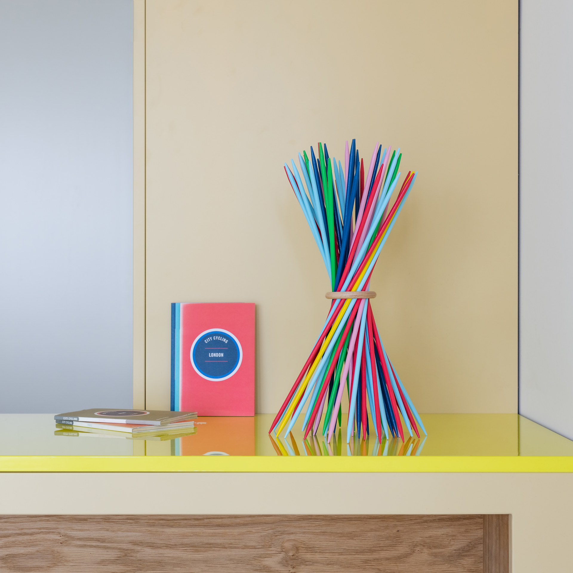 Kleurrijke houten stokjes, samengebonden door een houten ring, staan op een gele tafel. Boeken liggen ernaast. De achtergrond is lichtgeel en wit. Stickado is gerangschikt op de tafel.