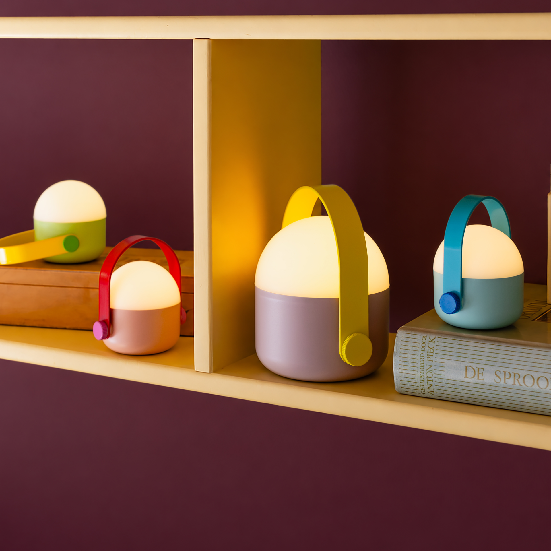 Drei akkubetriebene LED-Leuchten mit bunten Griffen stehen auf einem Regal: grün/gelb auf einer Holzbox, rosa/gelb daneben und rechts auf einem Buch hellblau mit türkisfarbenem Henkel. Alle leuchten warmweiß.