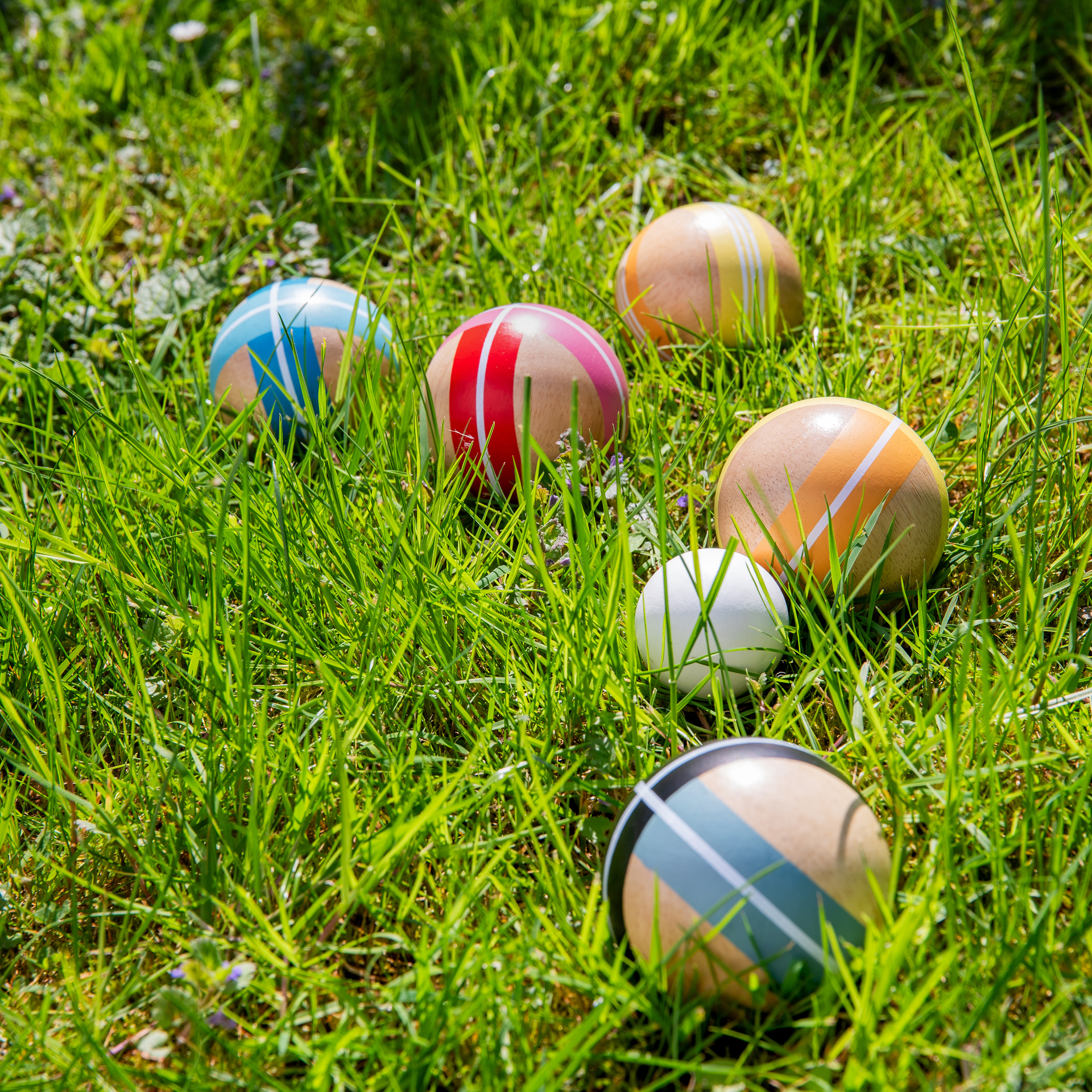 Bolas de madera de colores para petanca/boccia esparcidas sobre un césped soleado de color verde claro. Las bolas grandes son de color natural con rayas en azul, rojo, naranja y gris oscuro. Una pequeña bola blanca de objetivo yace en la hierba.