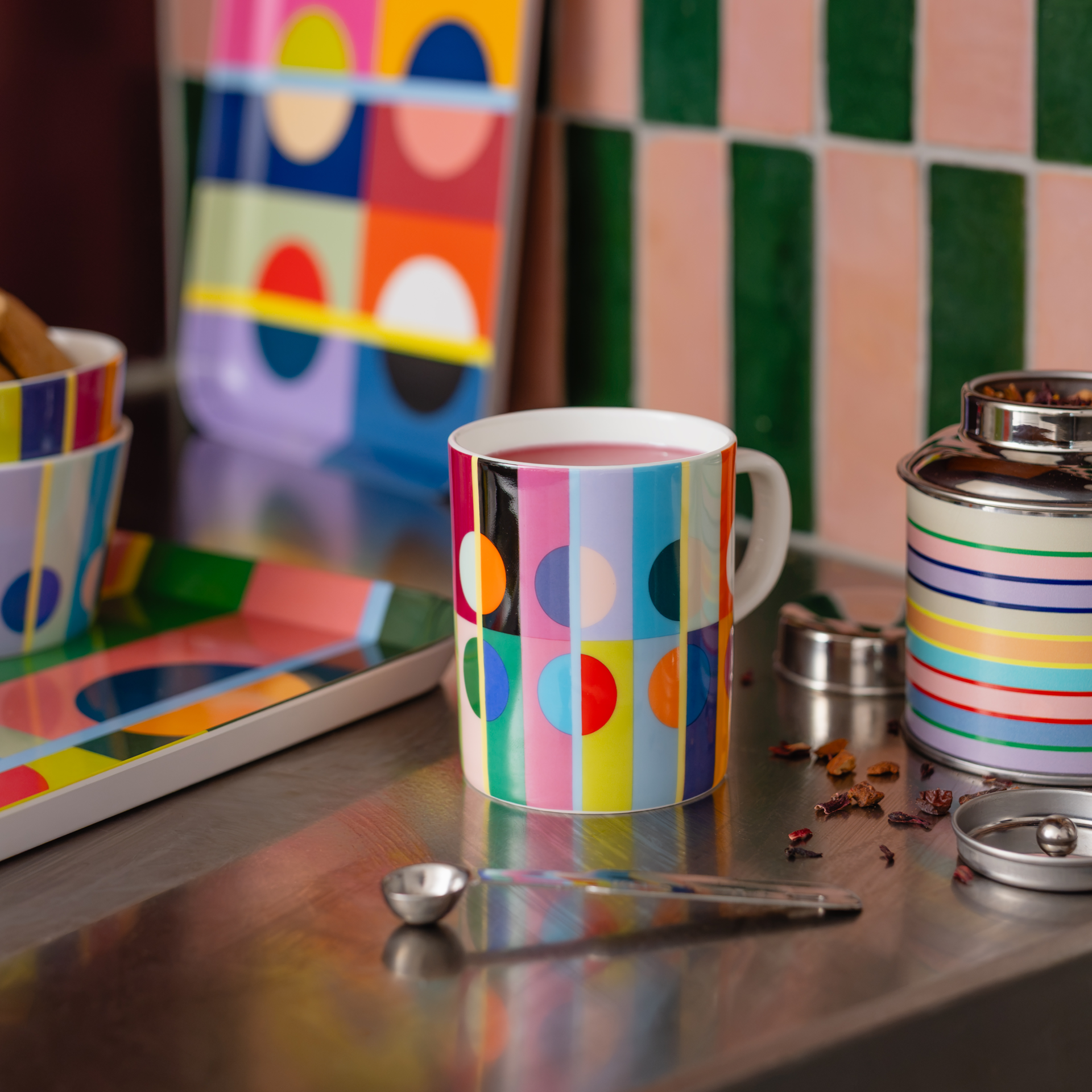 Taza de colores con rayas verticales y círculos, llena de una bebida rojiza, sobre una superficie metálica brillante. Junto a ella, una lata de té a rayas, hojas de té y una cucharita. Al fondo, vajilla de colores y una pared de azulejos verdes y rosas.
