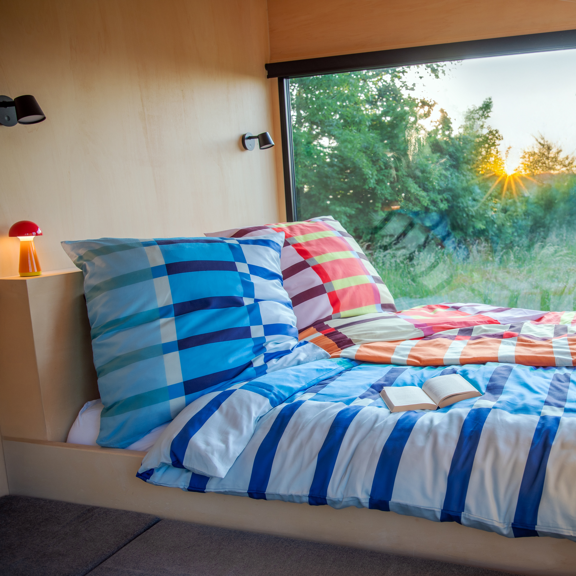 Gemütliches Bett mit Mako-Satin Bettwäsche 'Lucca' in blau/orange mit Streifen und Karomuster, ein offenes Buch liegt auf der Decke. Im Hintergrund ein Fenster mit Blick ins Grüne.