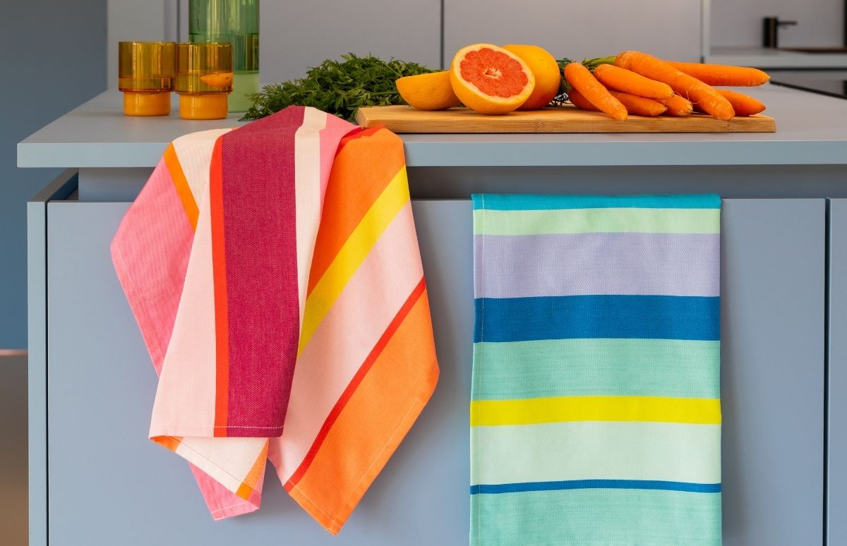 Zwei bunte, gestreifte Küchentücher hängen an hellblauen Schranktüren: links warme Streifen in Pink, Fuchsia, Orange, Gelb; rechts kühle in Blau, Lila, Grün. Auf der Arbeitsplatte liegen Karotten und Grapefruit auf Holzbrett.