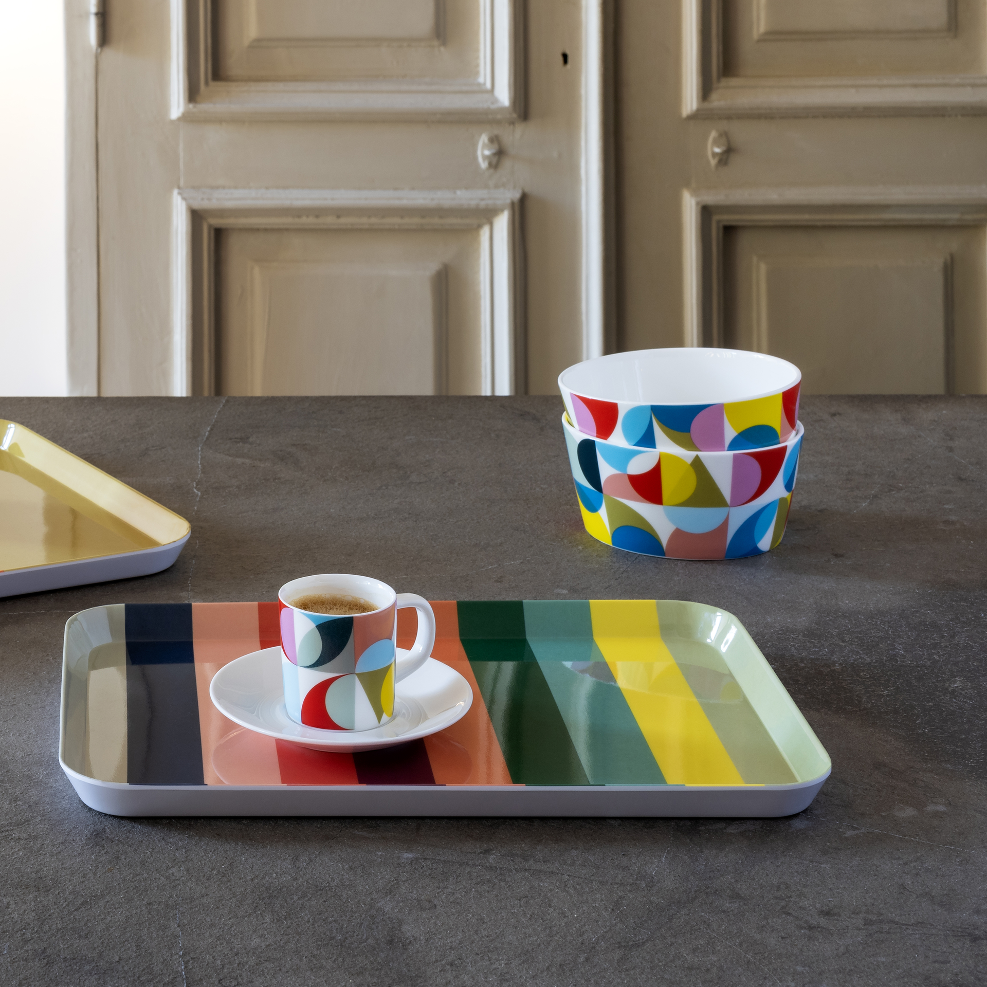 Due ciotole per cereali impilate con motivo geometrico, più una tazza da caffè espresso abbinata su un piattino, su un vassoio a righe colorate. Un altro vassoio è visibile ritagliato nell'immagine.