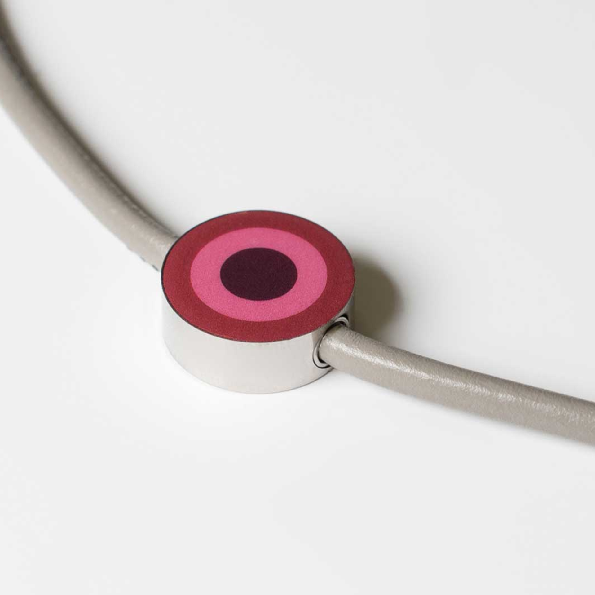 Gros plan sur le pendentif-bijou 13 avec un design circulaire dans les tons rose et lilas, relié à un ruban taupe. Le pendentif sert de fermeture.