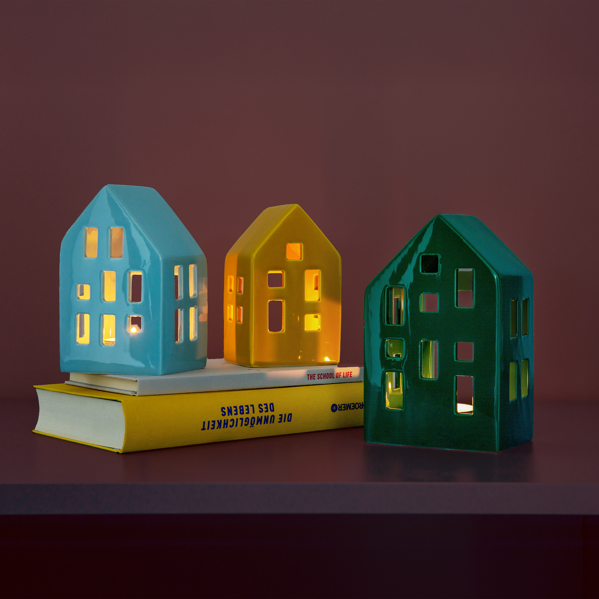 Three colourful ceramic house-shaped tealight holders on two books. The houses are blue, yellow and green. Candlelight is visible in the windows.