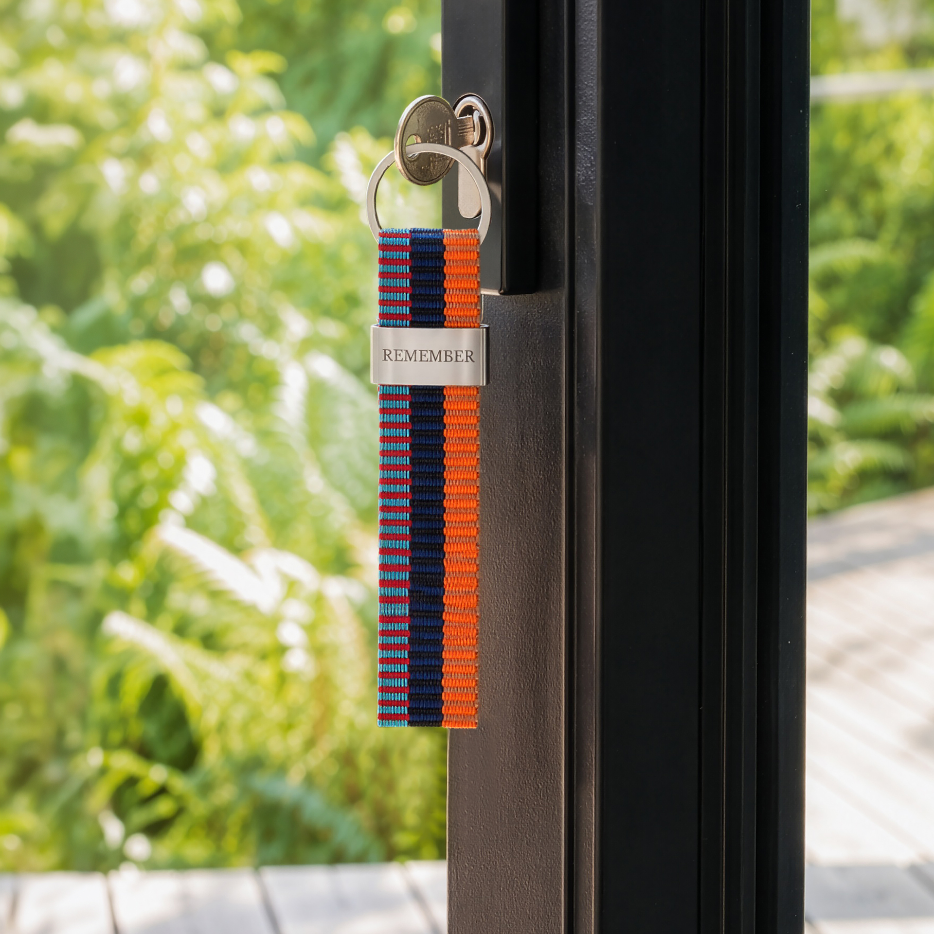 Längliches Schlüsselband mit farbigem Streifenmuster: vertikale Abschnitte in Rot, Hellblau, Dunkelblau und Orange mit feinen horizontalen Linien. Mittig silberne Metallplakette "REMEMBER", oben runder Schlüsselring.