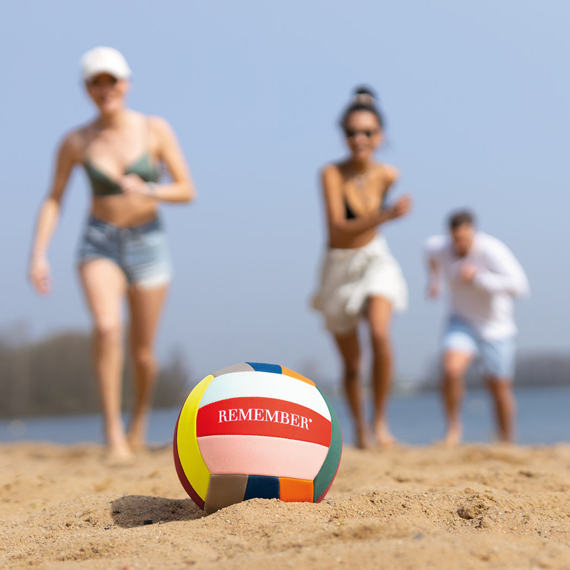 A coloured leisure ball lies on sand. Three people can be seen out of focus in the background. The ball is labelled "REMEMBER".