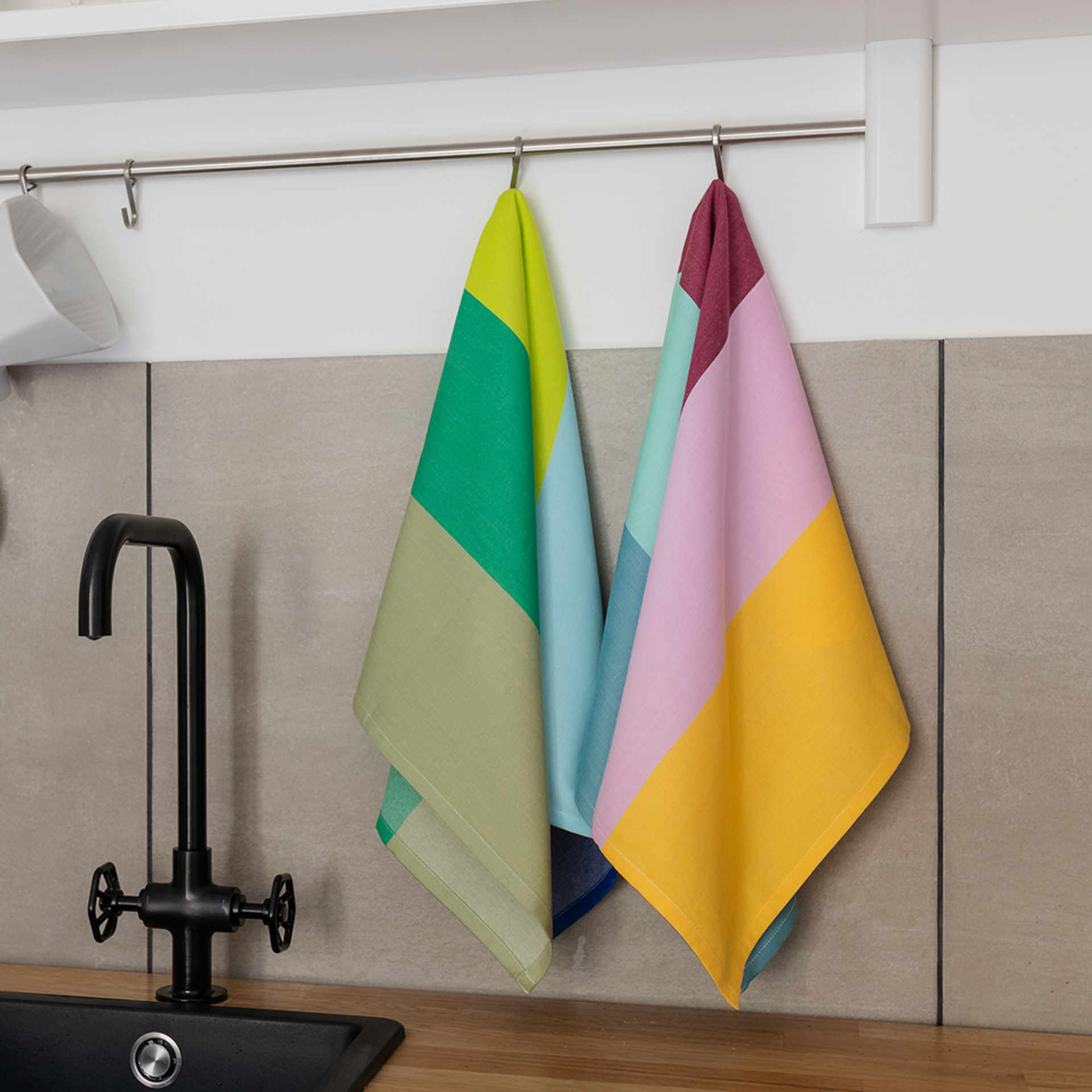 Deux sets de torchons aux motifs géométriques sont suspendus à un crochet devant un évier de cuisine. La serviette de gauche est dans des tons verts et bleus, celle de droite dans des tons jaunes et roses.
