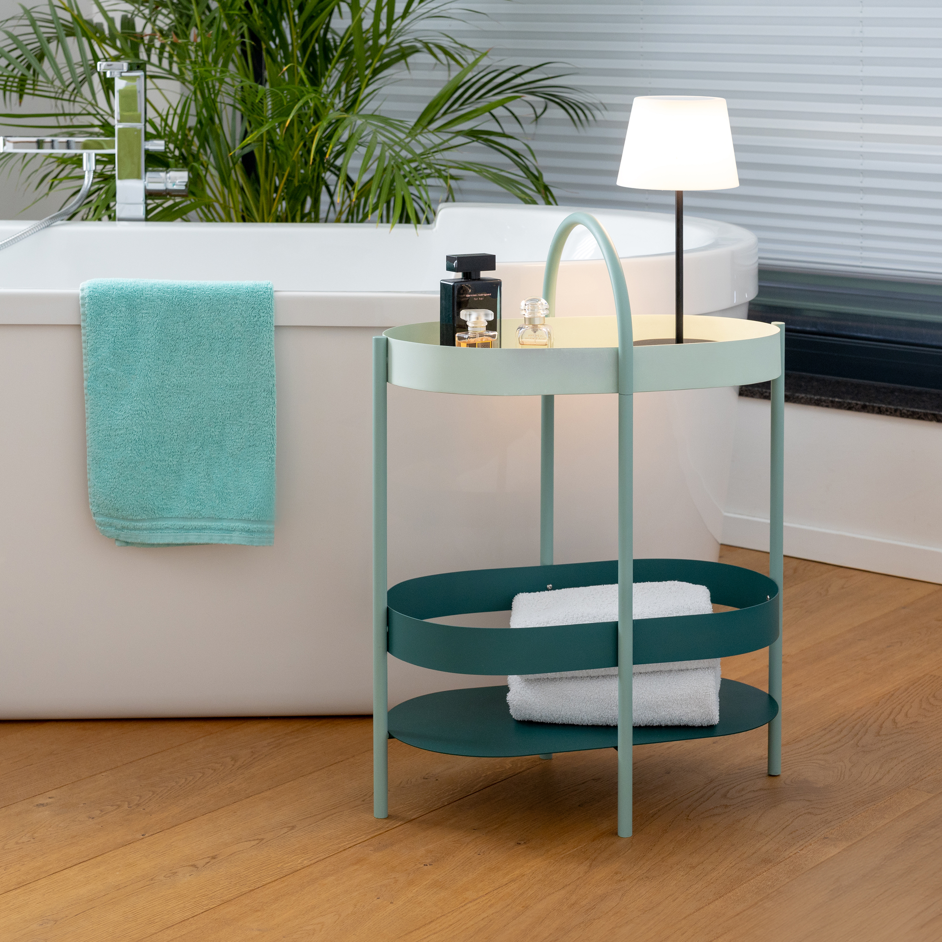 Eine grüne Beistellkonsole Barbara mit zwei Ablagen, Handtüchern und einer Lampe steht neben einer weißen Badewanne und einer grünen Pflanze auf einem Holzboden.
