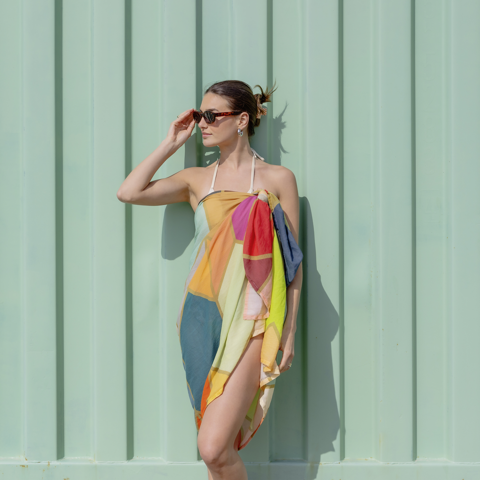 Mujer con gafas de sol que lleva un pareo Waikiki de colores a modo de vestido delante de una pared verde menta.