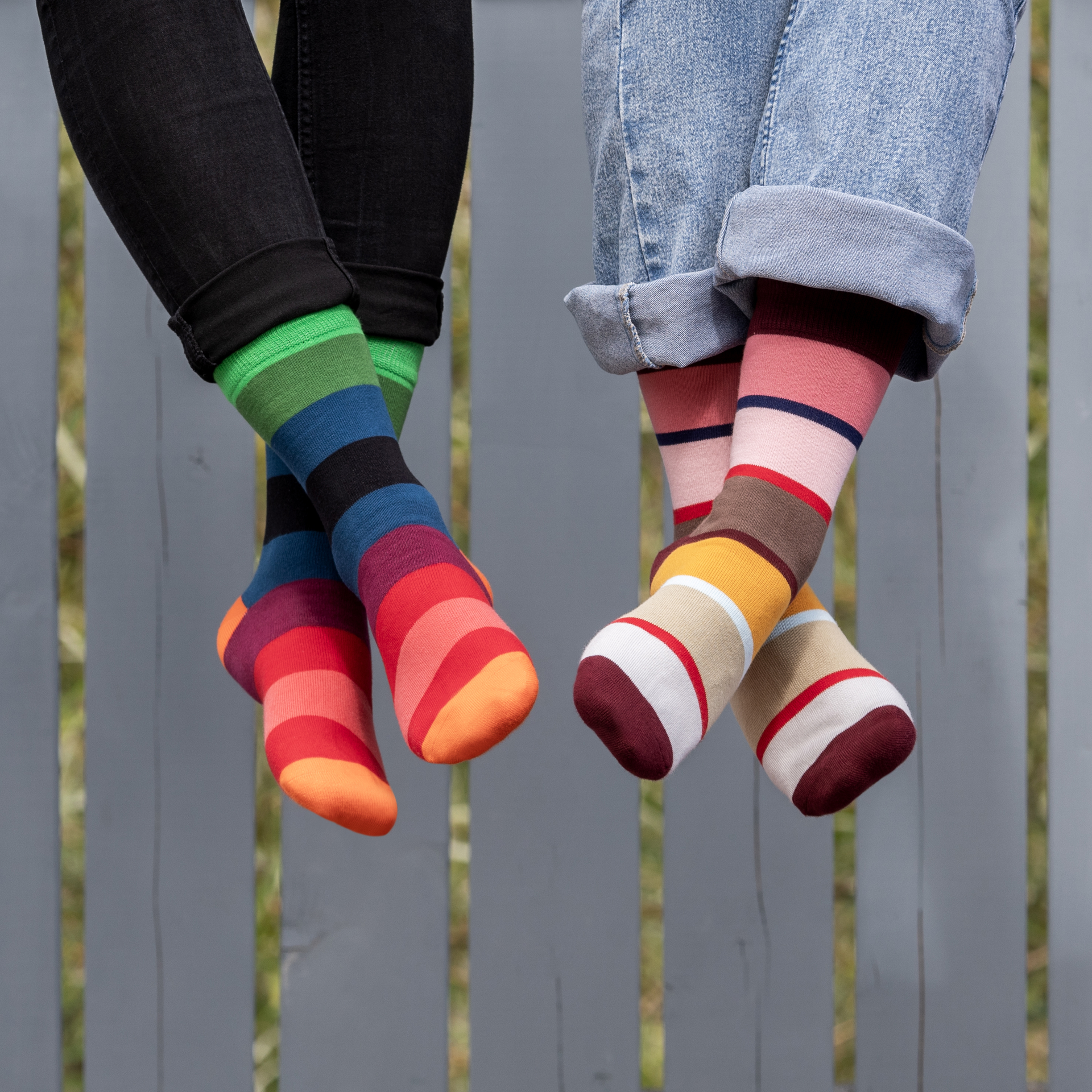 Dos pares de calcetines de diseño multicolor con rayas en las piernas sobre un fondo gris. A la izquierda, calcetines en tonos verdes, azules, rojos y naranjas; a la derecha, en tonos rojos, marrones, beige y blancos.