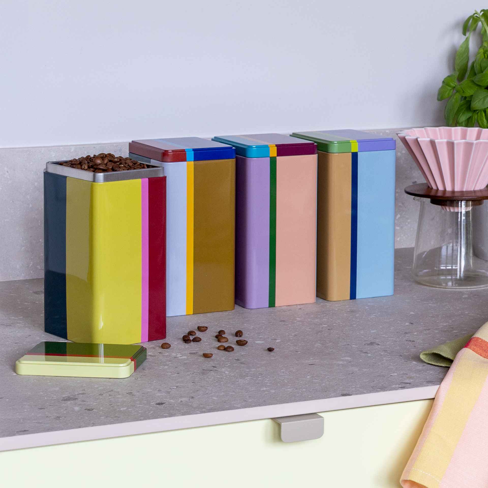 Vier bunte 'Yara' Dosen-Set, rechteckig, auf einer Arbeitsfläche. Eine Dose ist offen und mit Kaffeebohnen gefüllt, daneben liegen verstreut Kaffeebohnen.