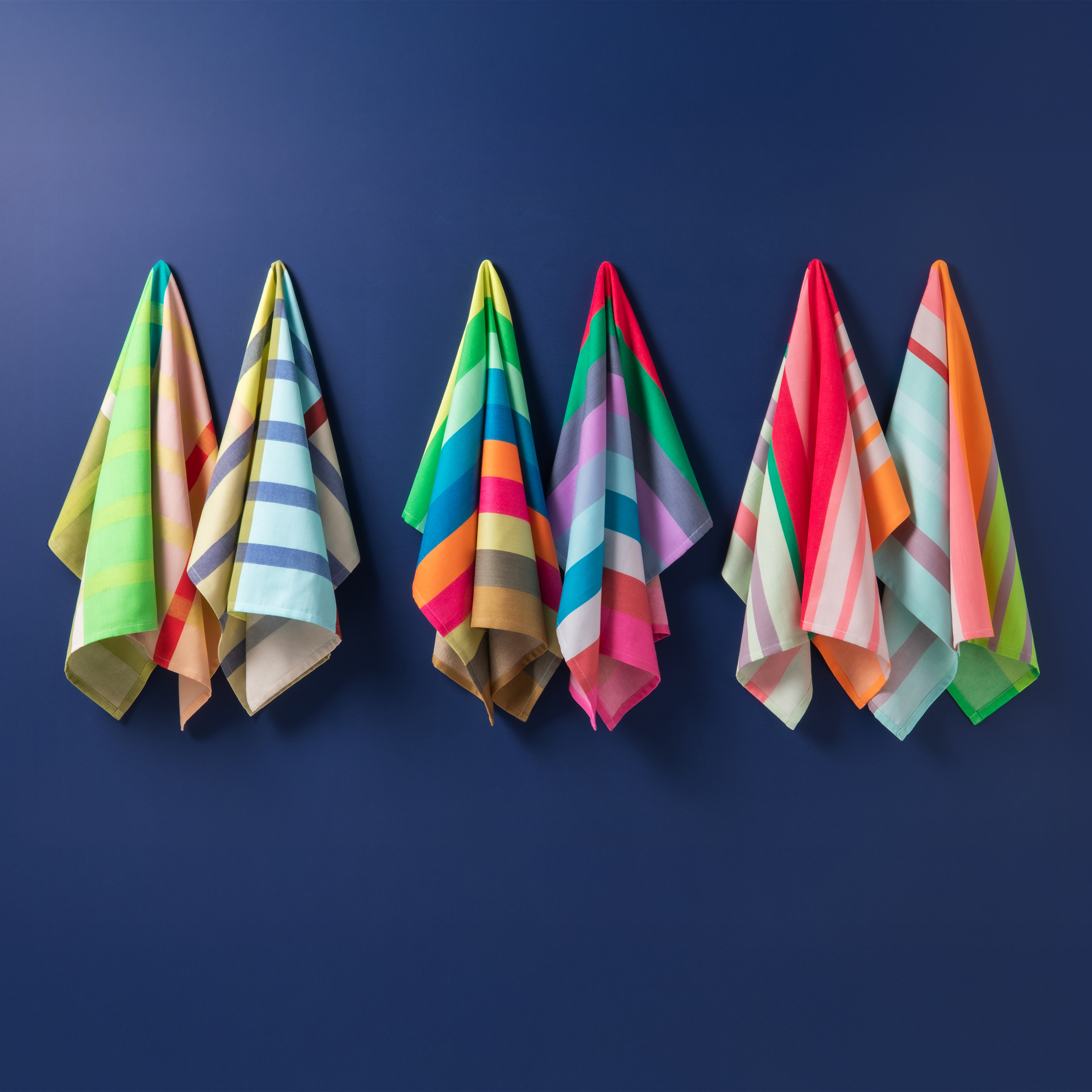 Six lingettes sèches rayées et colorées sont accrochées à un mur bleu foncé. Les lingettes sont disposées par paires et présentent différentes combinaisons de couleurs.