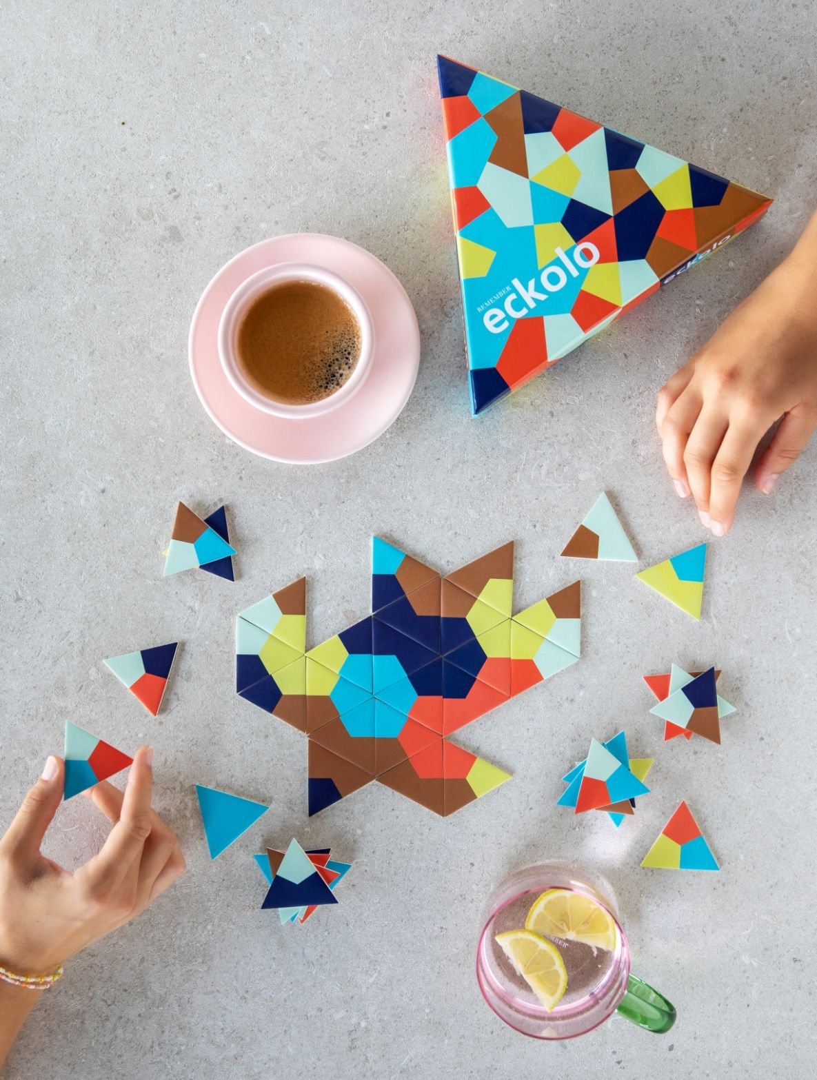 Geometrisches Puzzle-Spiel Eckolo auf einem Tisch. Große sternförmige Anordnung bunter Dreiecke (Braun, Blau, Grün, Orange) in der Mitte. Zwei Hände legen Steine. Daneben rosa Kaffeetasse, Wasser mit Zitrone, Box sichtbar.
