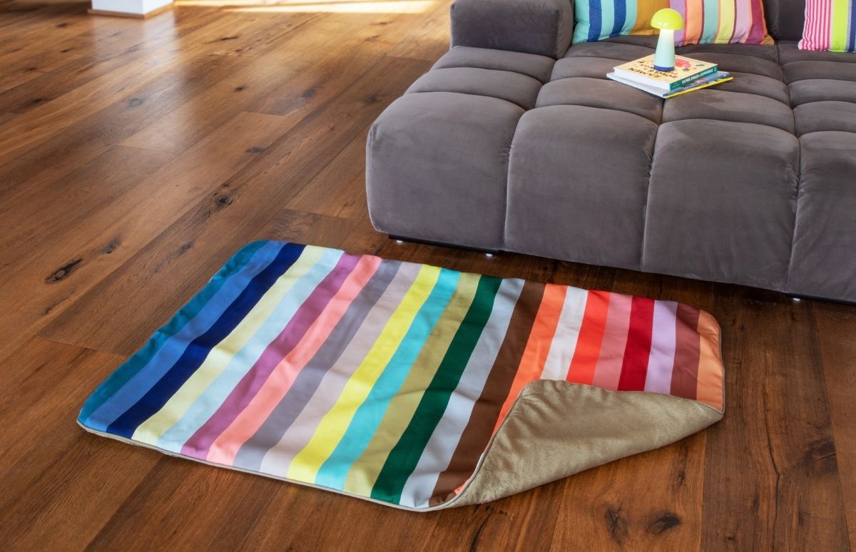 Eine farbenfrohe, gestreifte Decke liegt auf warmem Holzboden vor einem grauen, modularen Sofa. Sie zeigt breite, vertikale Streifen in Blau, Grün, Gelb, Orange, Rot und Pastelltönen. Auf dem Sofa liegen bunte Kissen.