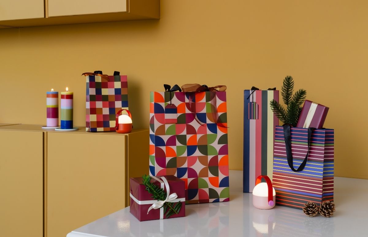 Festliche Anordnung bunter Geschenktüten und Päckchen auf weißem Tisch vor senfgelber Wand. Tüten zeigen lebendige Karos, Halbkreise und Streifen. Dazu brennen gestreifte Kerzen und leuchten kleine Portable-Lampen, umgeben von Tannenzweigen und Zapfen.