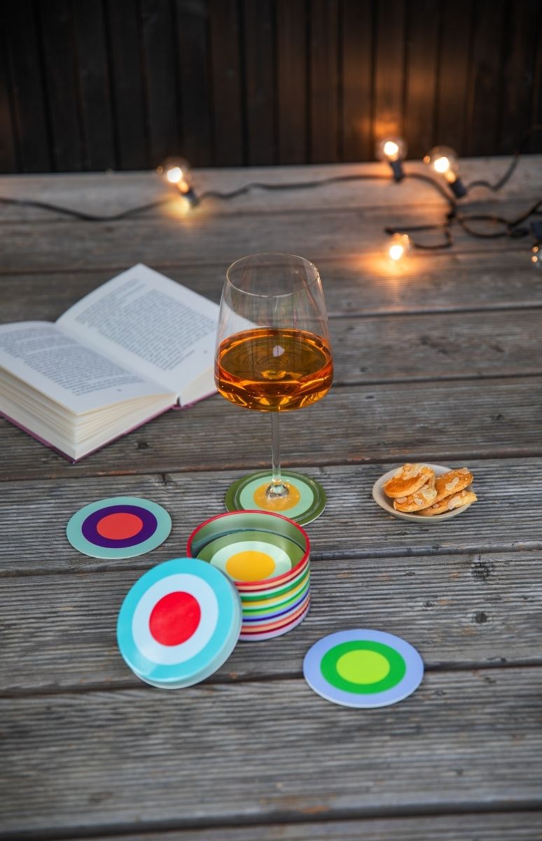 Auf einem Holztisch liegen ein offenes Buch, ein Weinglas mit bernsteinfarbenem Getränk auf buntem Untersetzer und ein Teller mit Mandelkeksen. Daneben farbenfrohe runde Untersetzer, teils im Behälter. Im Hintergrund Lichterketten.