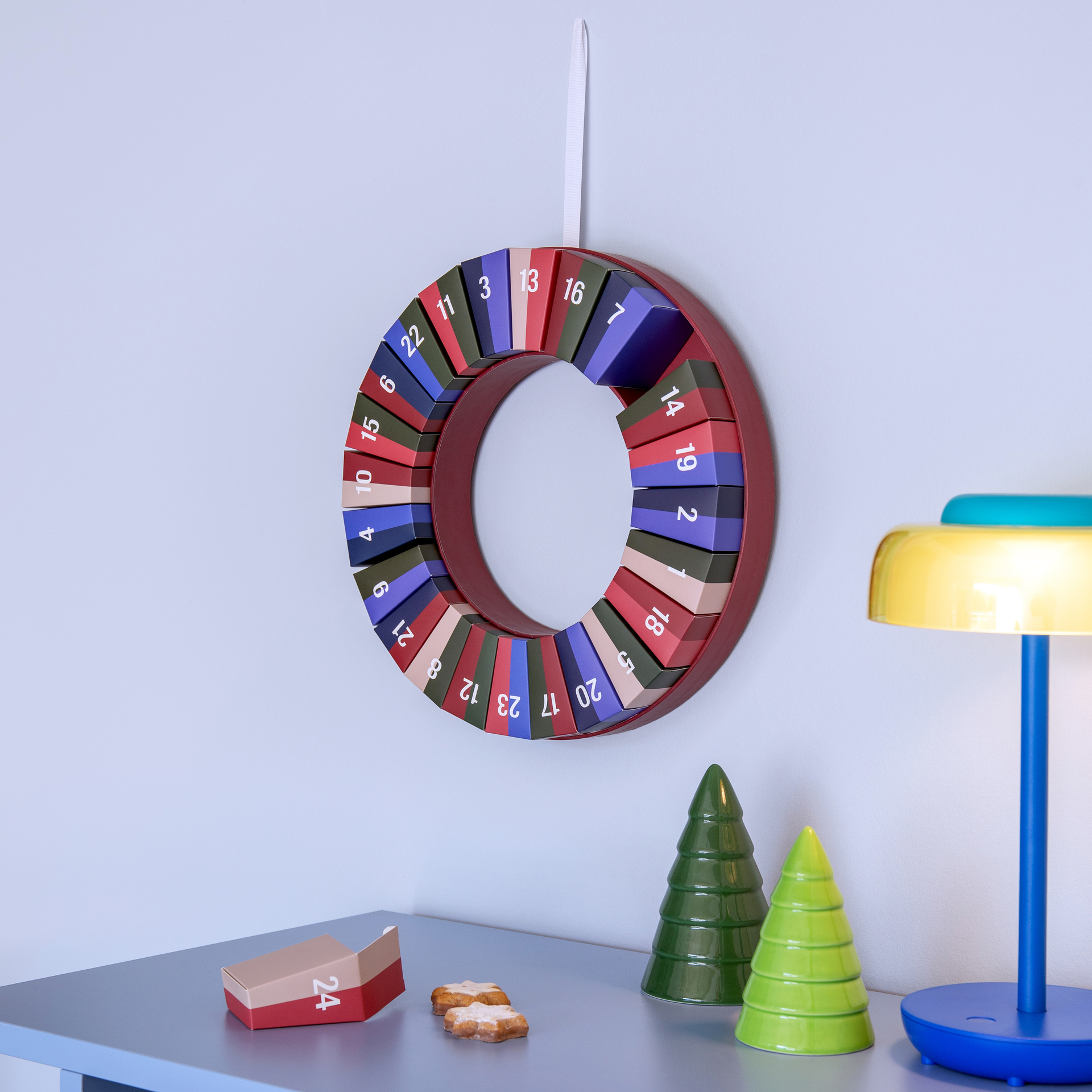 Corona de Adviento moderna redonda en pared azul claro, con 24 cajitas de colores numeradas en rojo, verde, azul y beige sobre aro rojo oscuro. En la mesa: dos galletas estrella, caja 24 abierta y dos abetos.