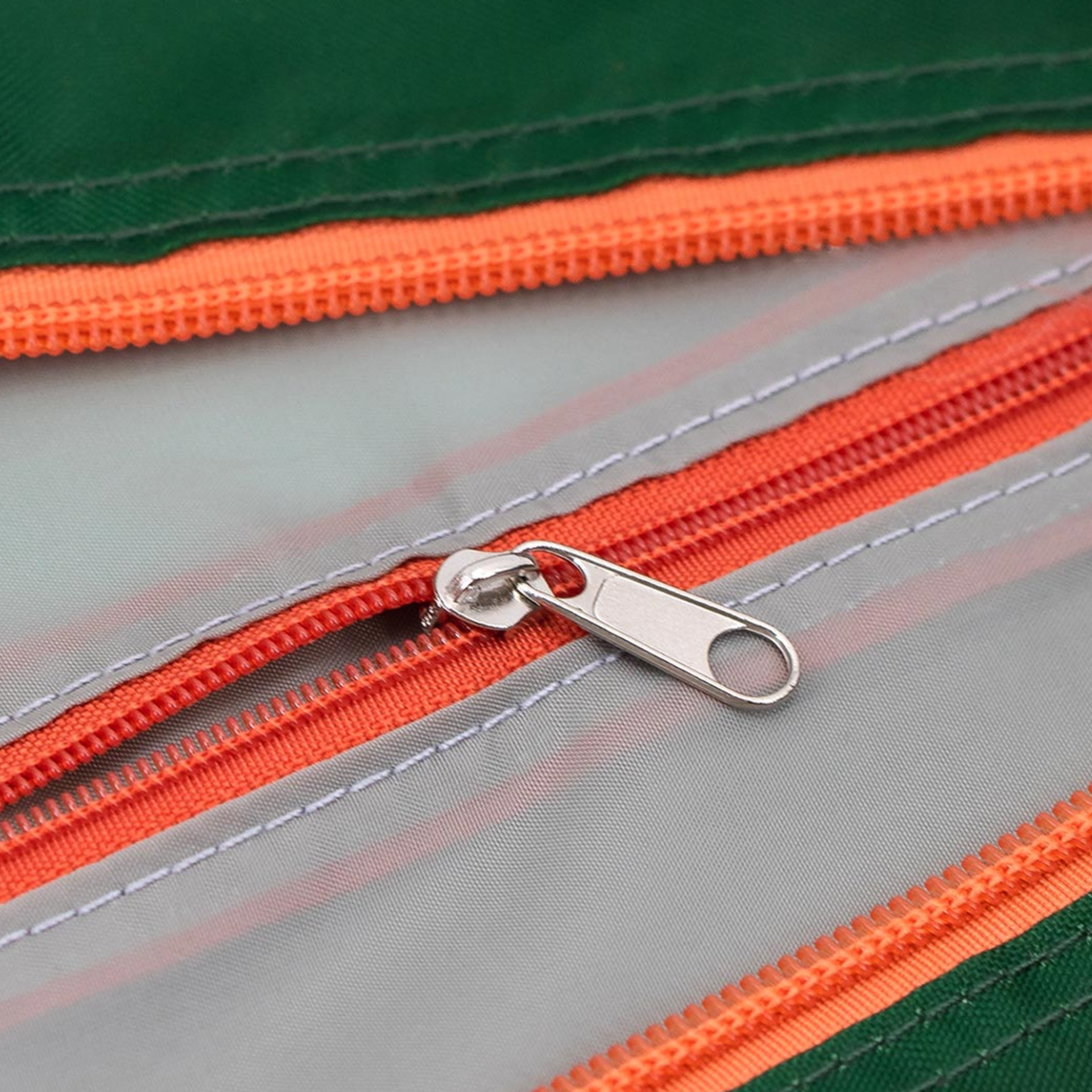 Detalle de una bolsa de viaje «Max»: la cremallera está abierta, la cremallera plateada descansa sobre la tela gris claro. Se pueden ver las costuras y las cremalleras de color naranja.