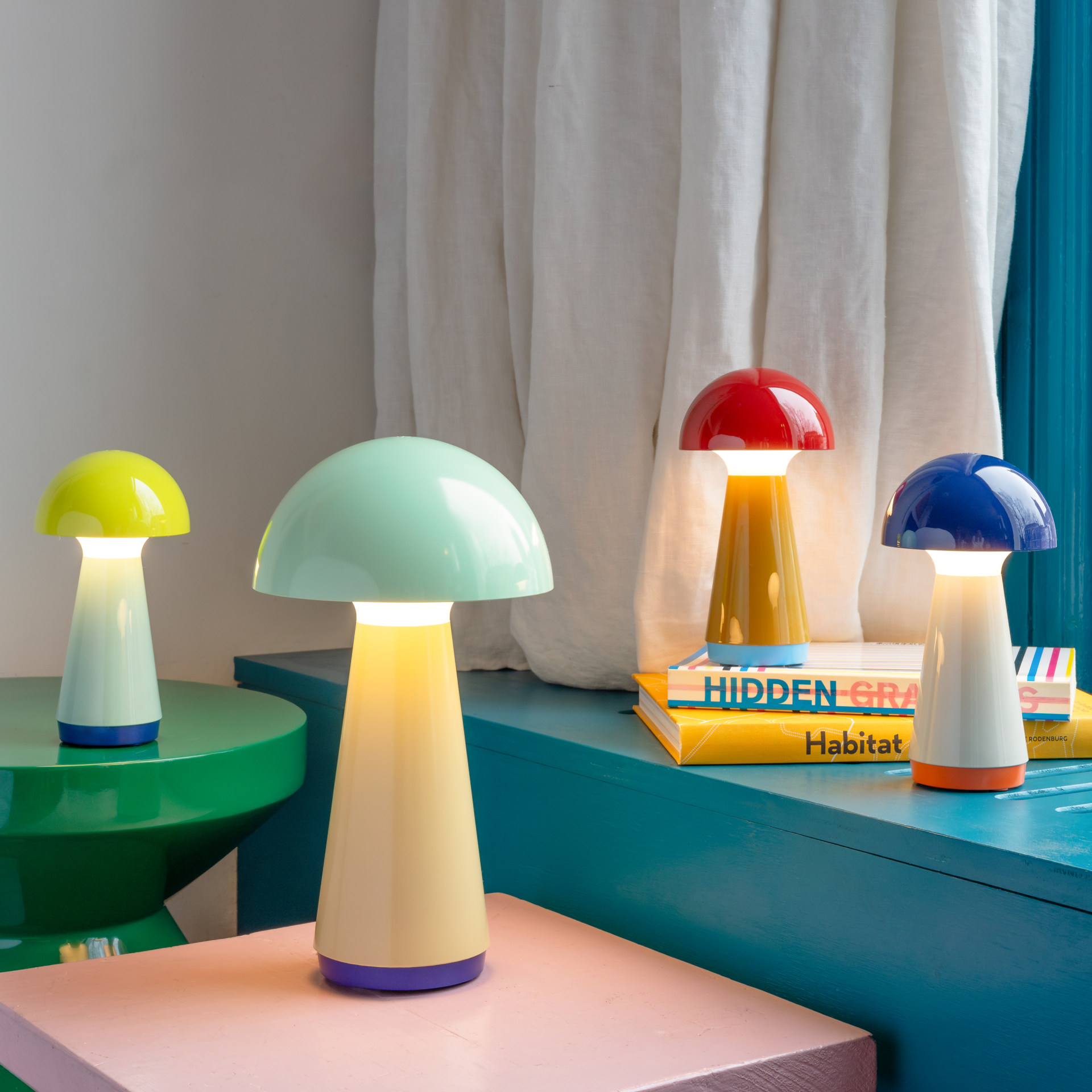 Cuatro lámparas de mesa con forma de seta «Bob» en colores menta, amarillo, rojo y azul iluminan mesas de colores y pilas de libros frente a unas cortinas y una pared.