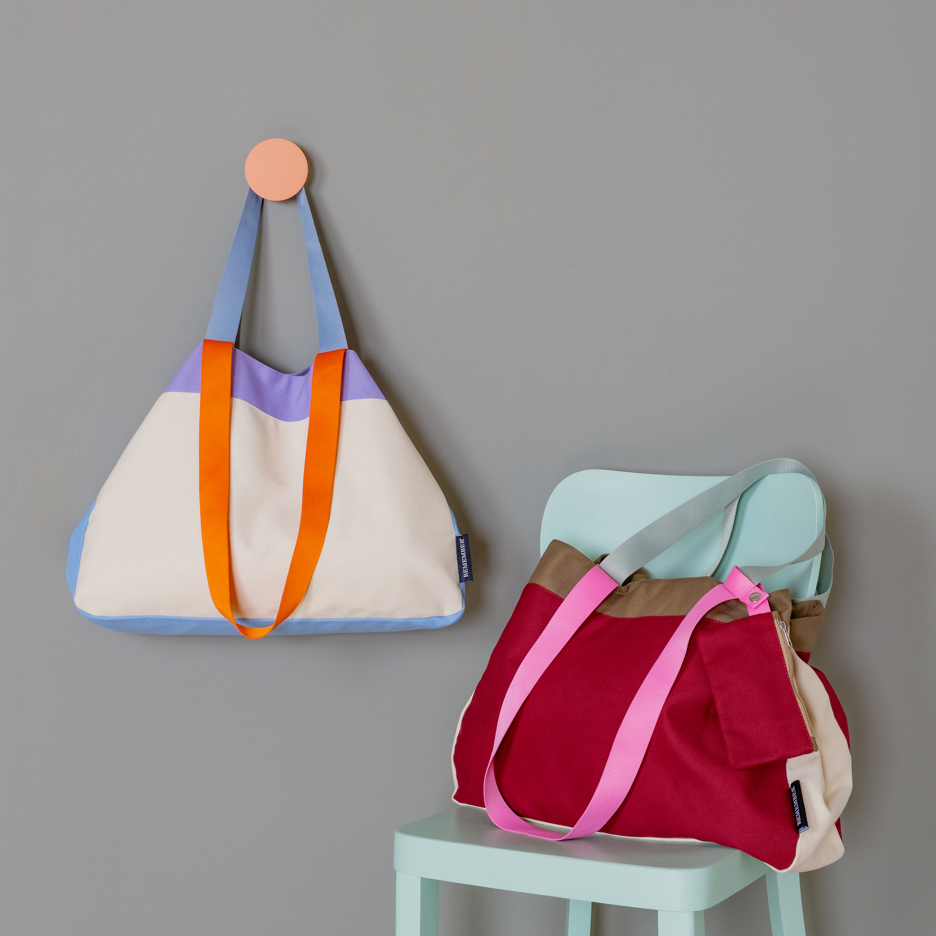 Zwei Canvas Taschen 'Emma' in verschiedenen Farben vor grauer Wand. Eine hängt an einem runden, farbigen Wandhaken, die andere steht auf einem Stuhl. Beide haben farbige Trageriemen.
