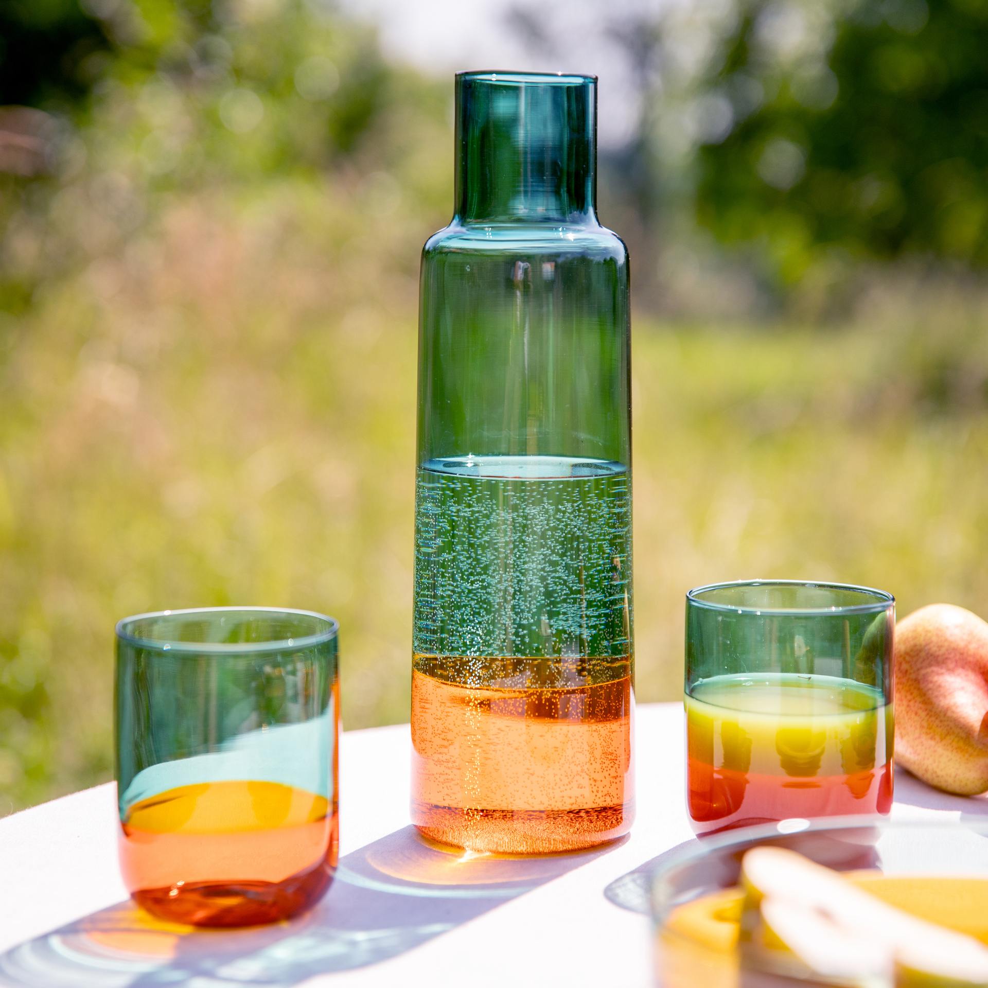 Una sottile caraffa di vetro 'ambra' riempita con un liquido gorgogliante, affiancata da due bicchieri coordinati su un tavolo all'aperto, lo sfondo è sfocato.