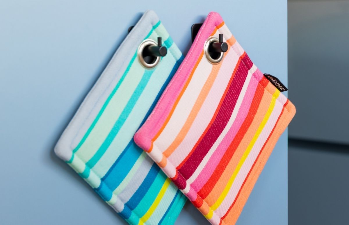 Zwei bunte, gestreifte Topflappen hängen an schwarzen Haken vor hellblauem Hintergrund. Links Streifen in Blau- und Türkistönen, rechts in Pink, Rot, Orange, Gelb und Weiß. Beide mit silberner Öse; rechts mit schwarzem Etikett.