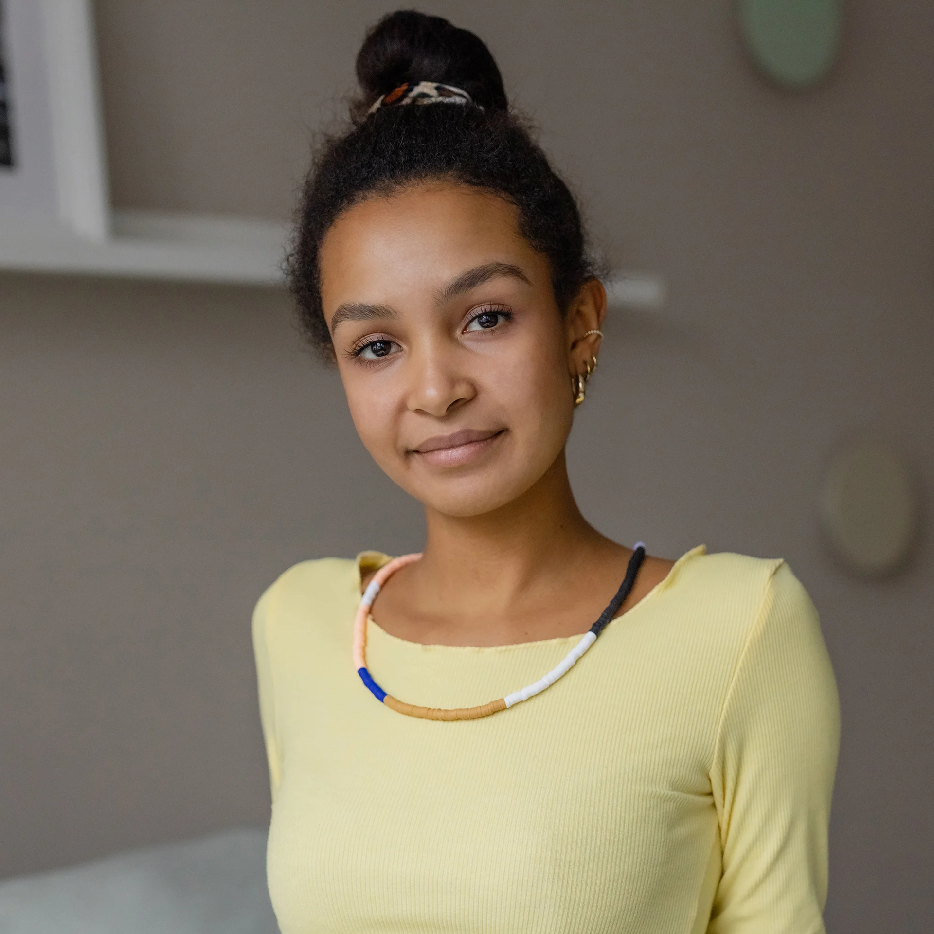 Een jonge vrouw met donkere huid en donker, krullend haar in een knot kijkt met een zachte glimlach naar de camera. Ze draagt een geribbeld lichtgeel shirt met lange mouwen, een kleurrijke kralenketting en gouden oorbellen.
