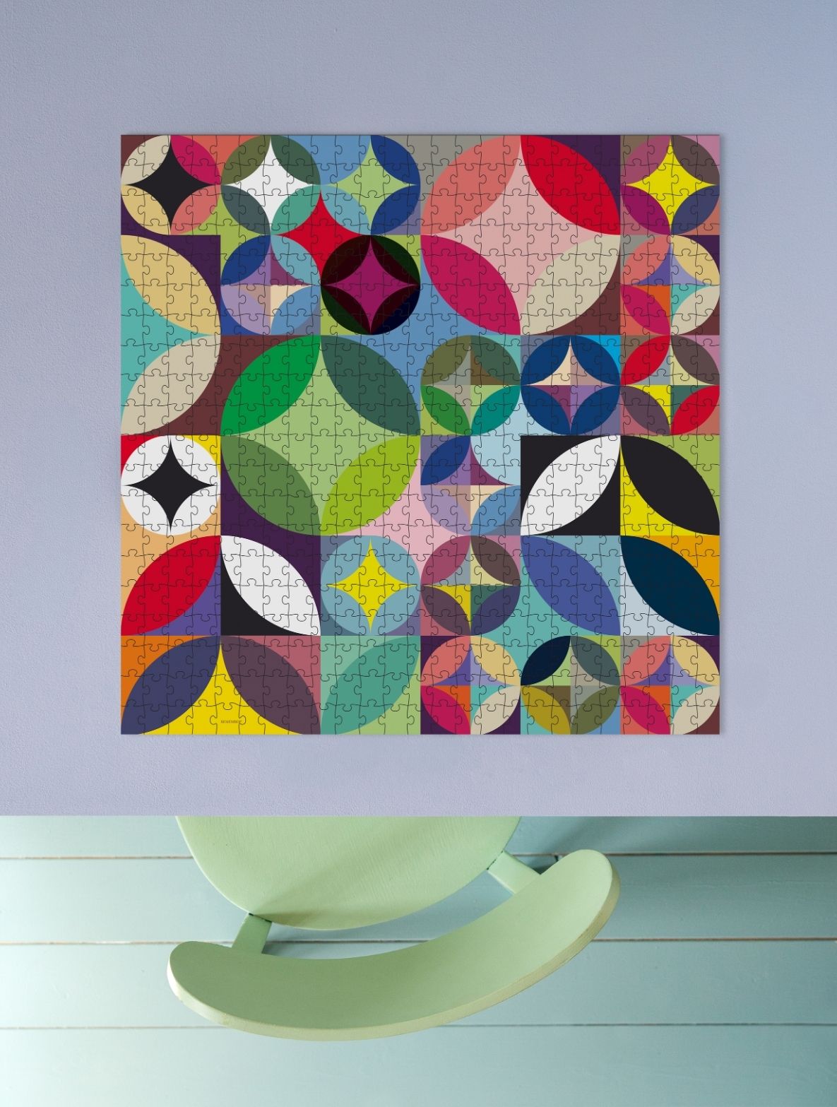 Ein farbenfrohes Jigsaw-Puzzle hängt an einer hellvioletten Wand. Es zeigt ein abstraktes Muster aus überlappenden Kreisen und blattartigen Formen in Rot, Blau, Grün, Gelb, Schwarz und Weiß. Darunter ein hellgrüner Schaukelstuhl.