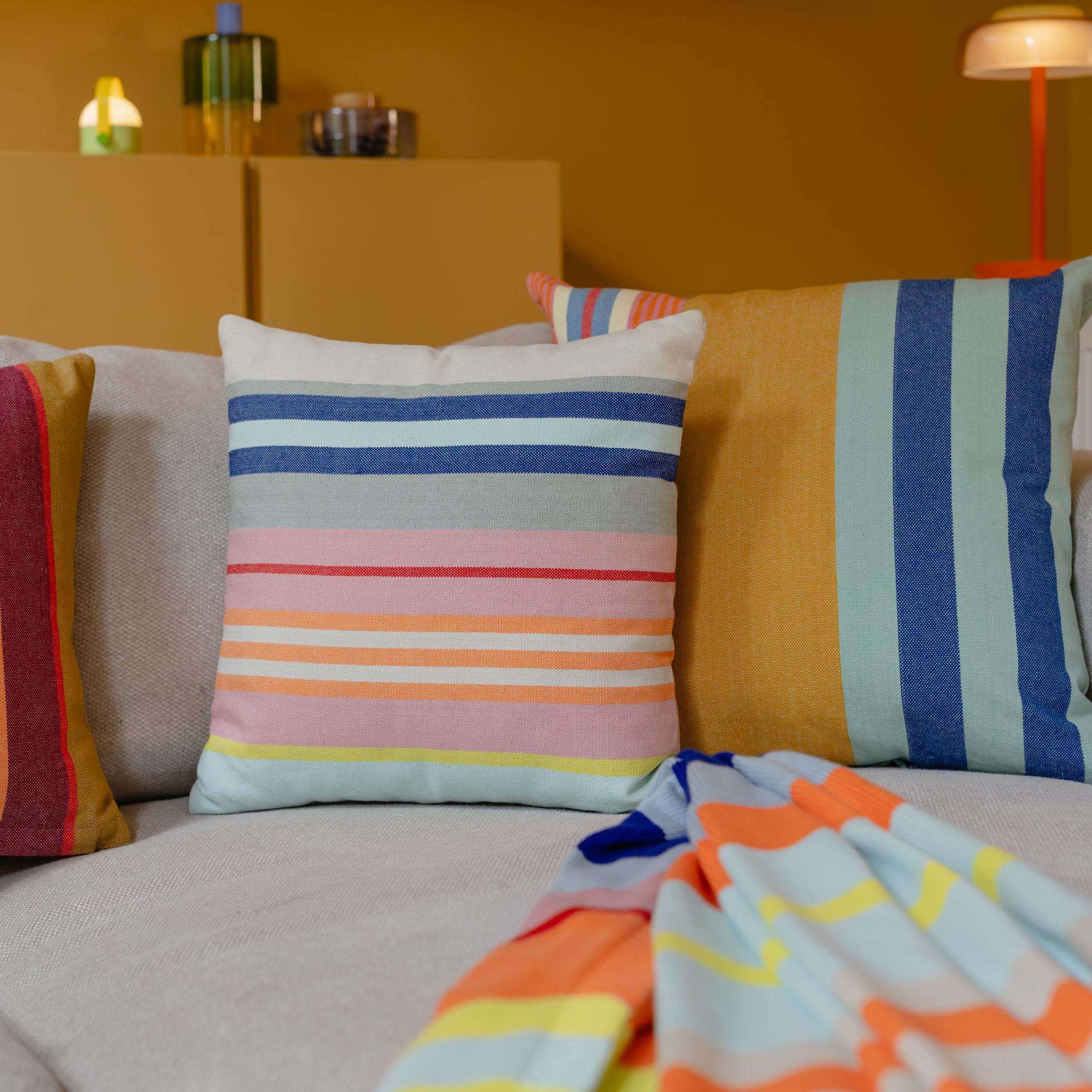 Quadratisches Baumwollkissen in sanften Sommerfarben. Horizontale Streifen in unterschiedlichen Breiten: Beige, Blau- und Grüntöne, Pink, Rot, leuchtendes Orange und Gelb. Grob gewebte Oberfläche sichtbar, Markenetikett unten rechts.