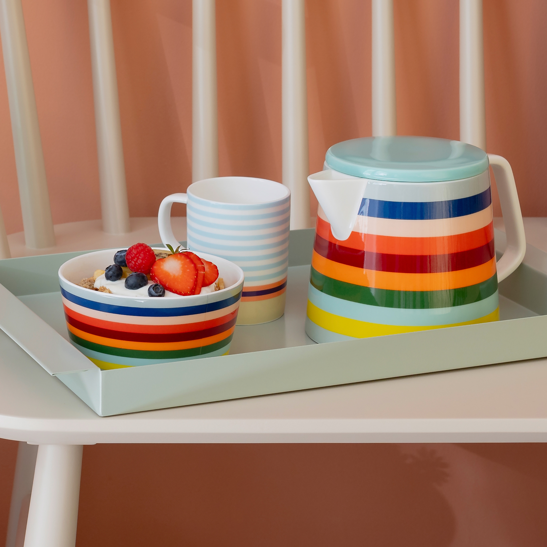 Una teiera "Lorenzo" a righe colorate, una tazza a righe azzurre e una ciotola di yogurt e frutta fresca su un vassoio verde menta.