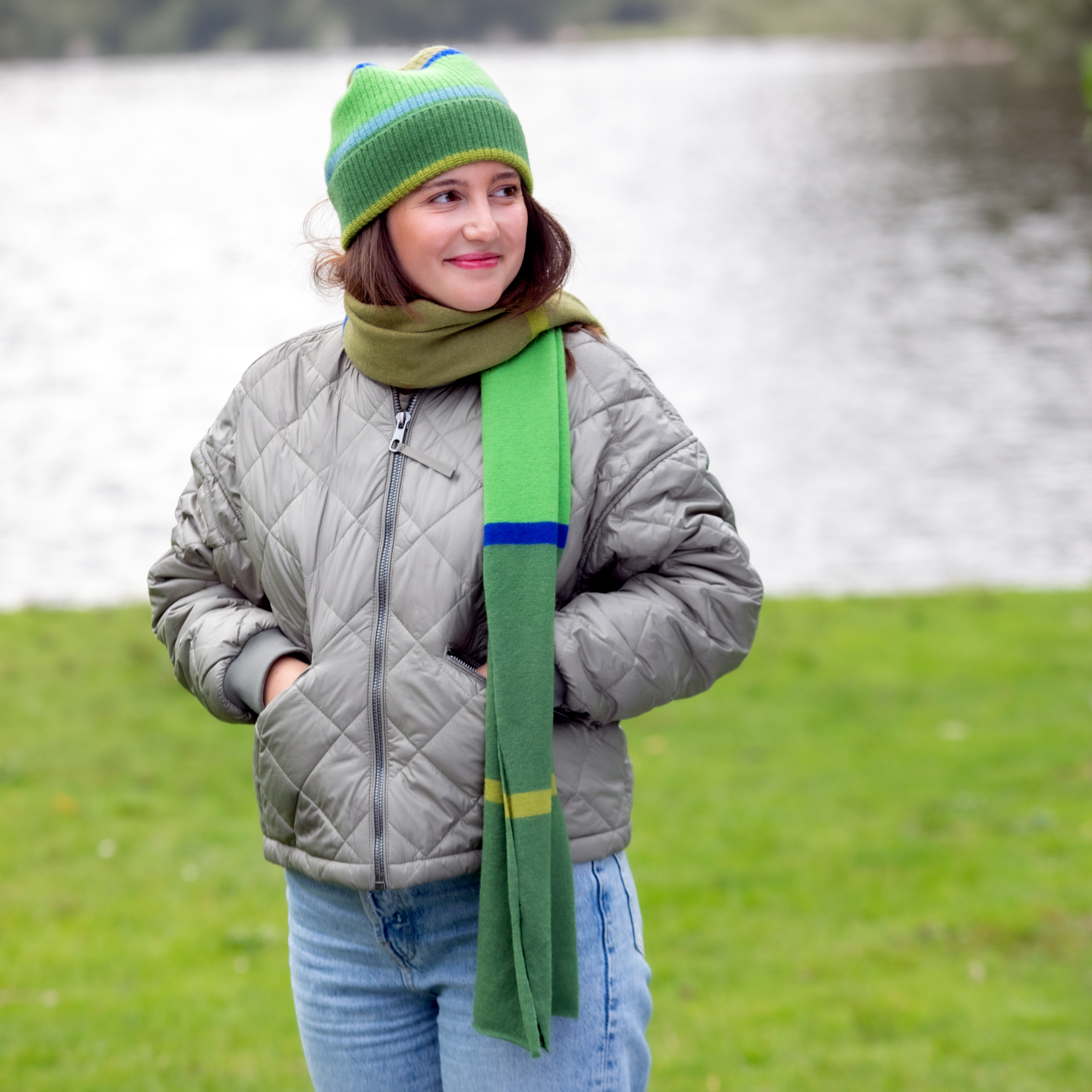 Una giovane donna con cappello e sciarpa verde è in piedi su un prato di fronte a un lago. Indossa una giacca trapuntata e dei jeans e guarda alla sua destra con un sorriso.