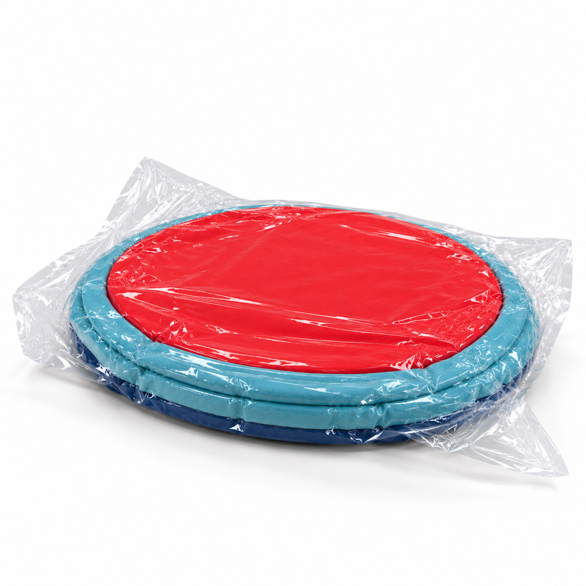Ein runder Pouf, dreifarbig in Rot, Hellblau und Dunkelblau, ist in transparenter Vakuumverpackung eingeschweißt. Der Pouf wirkt komprimiert und flach, bereit zum Auspacken und Entfalten. Die einzelnen Farbschichten sind deutlich erkennbar.