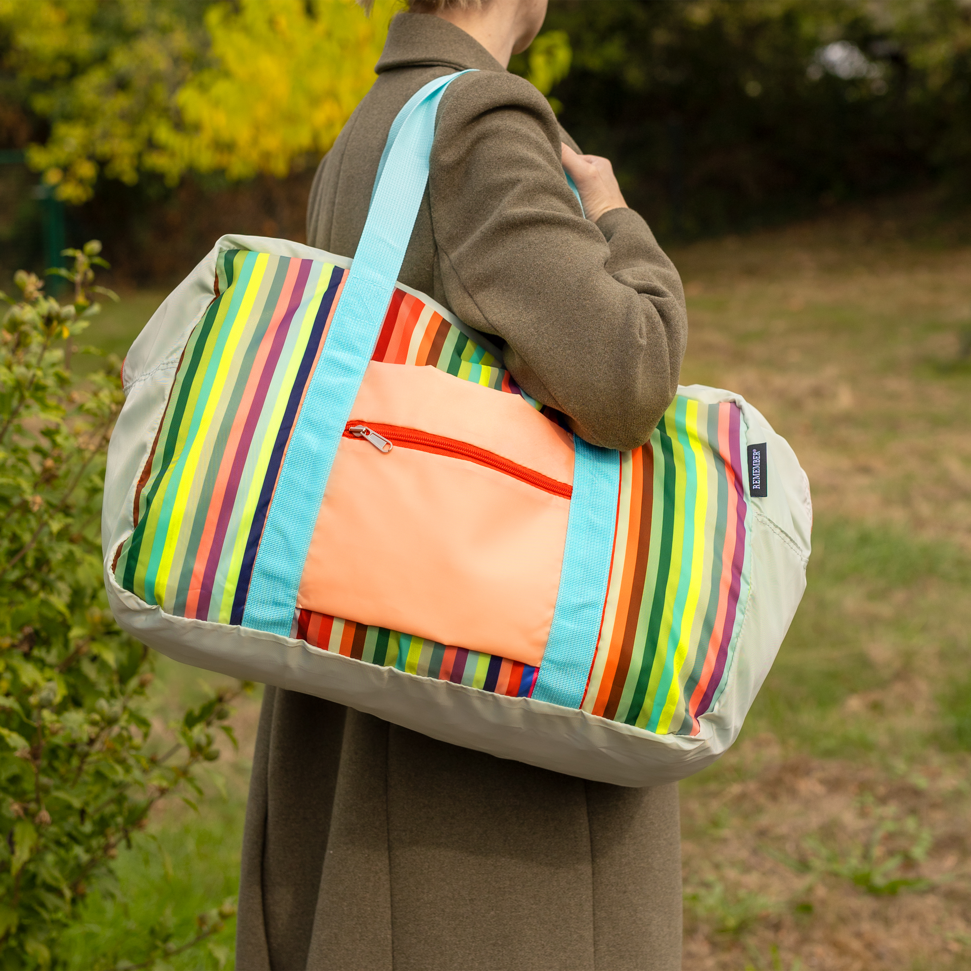 Una persona lleva al hombro una bolsa de ocio y viaje con rayas verticales rojas, verdes, azules y amarillas. De poliéster resistente, con correas azul claro y bolsillo exterior naranja con cremallera. Ideal para escapadas o piscina.