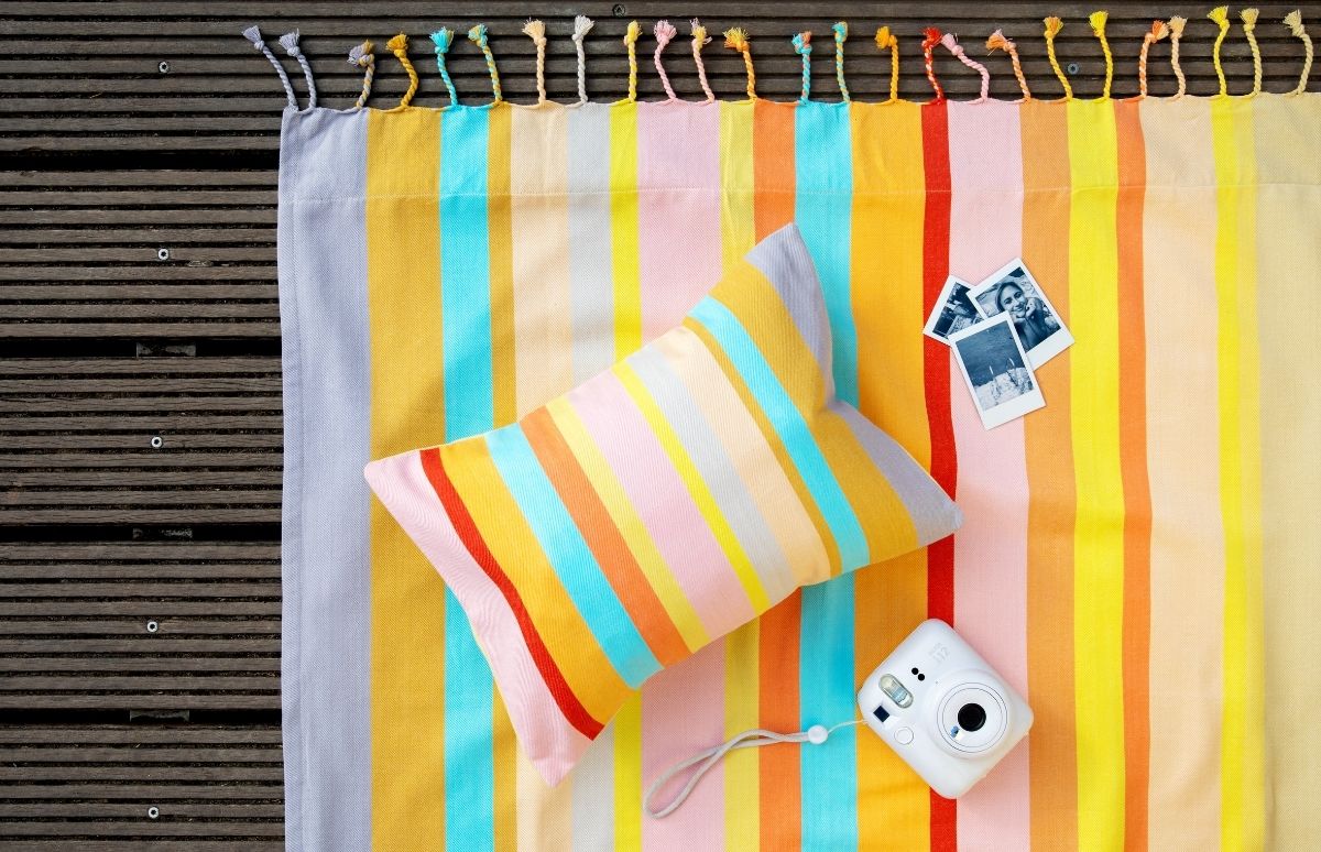 Bunte, gestreifte Stranddecke mit Quasten auf dunklen Holzplanken. Darauf ein passendes Kissen in Gelb, Orange, Pink und Blau sowie eine weiße Sofortbildkamera. Daneben drei kleine S/W-Polaroids mit Personen und Strand.