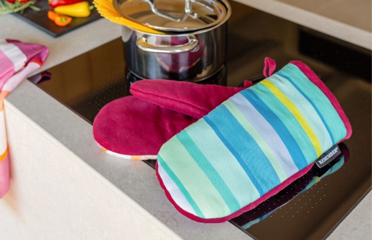 Auf schwarzer Induktionskochplatte liegen zwei bunte Ofenhandschuhe neben einem Edelstahltopf mit Spaghetti. Einer ist magenta, der andere hat Querstreifen in Türkis, Grün, Gelb und Blau mit pinkem Rand. Unscharf: Paprika und Kräuter auf Schneidebrett.