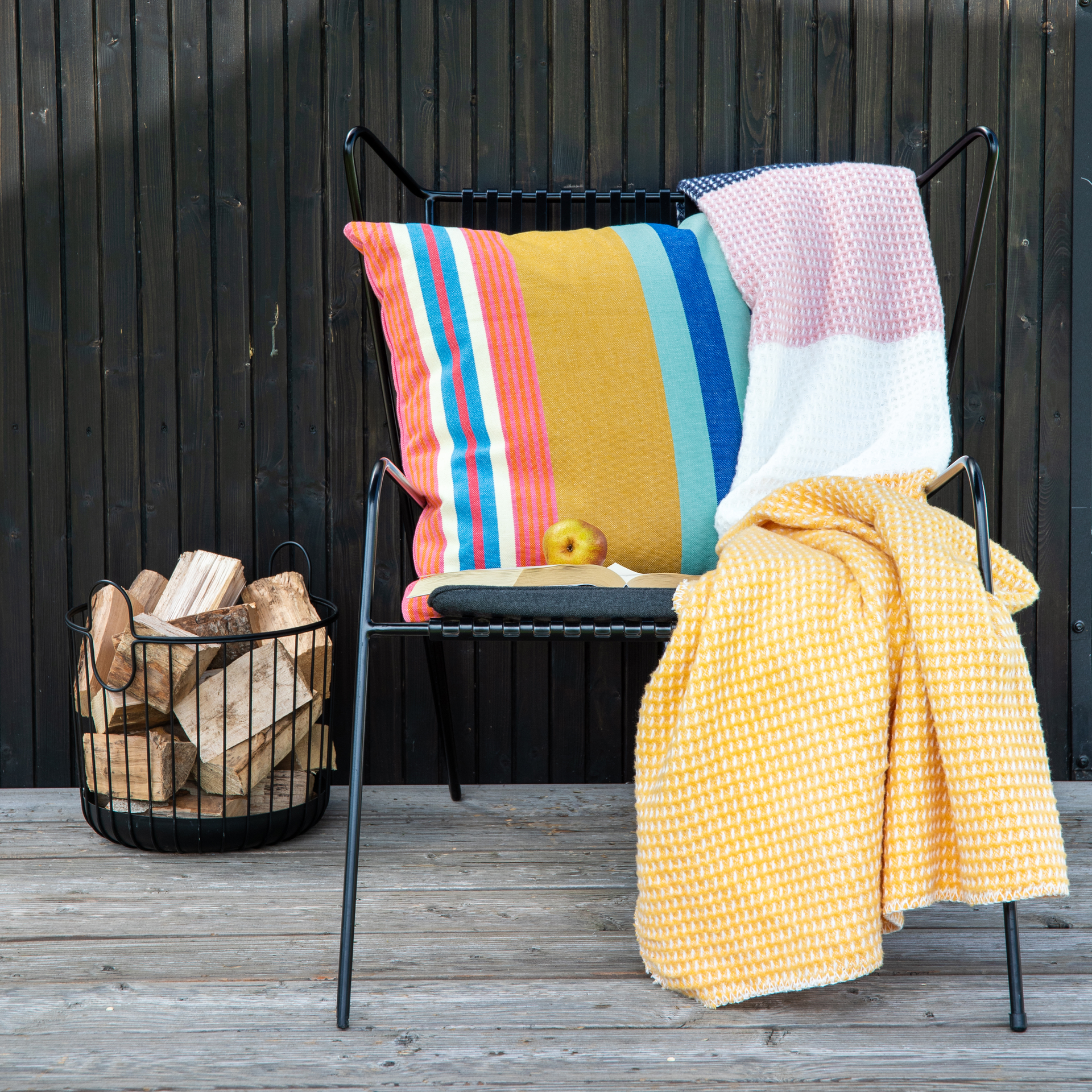 Sedia in metallo nero con cuscini in cotone "Belize" a righe, coperte, mele e libri davanti a una parete in legno scuro e a un cesto di legna.