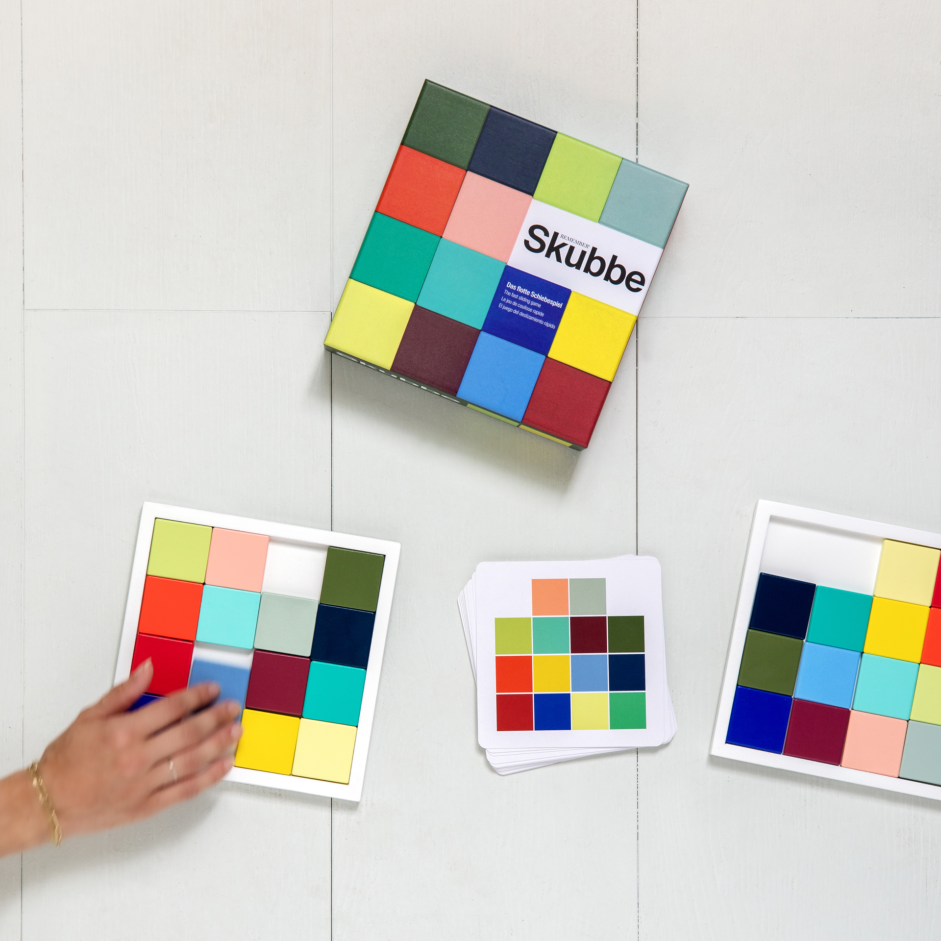 Vista superior del juego «Skubbe»: tablero con fichas de madera de colores, cartas de referencia y caja del juego sobre fondo blanco. Una mano empuja una ficha.