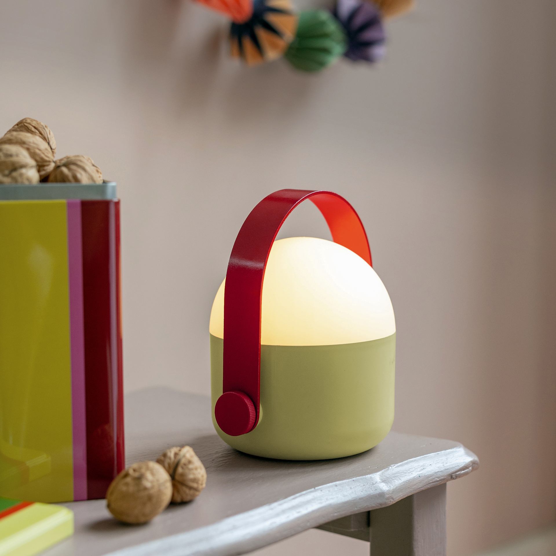 Verlicht, zandkleurig LED-lampje met rood handvat. Het staat op een grijze tafel. Een kleurrijk blik noten staat ernaast.