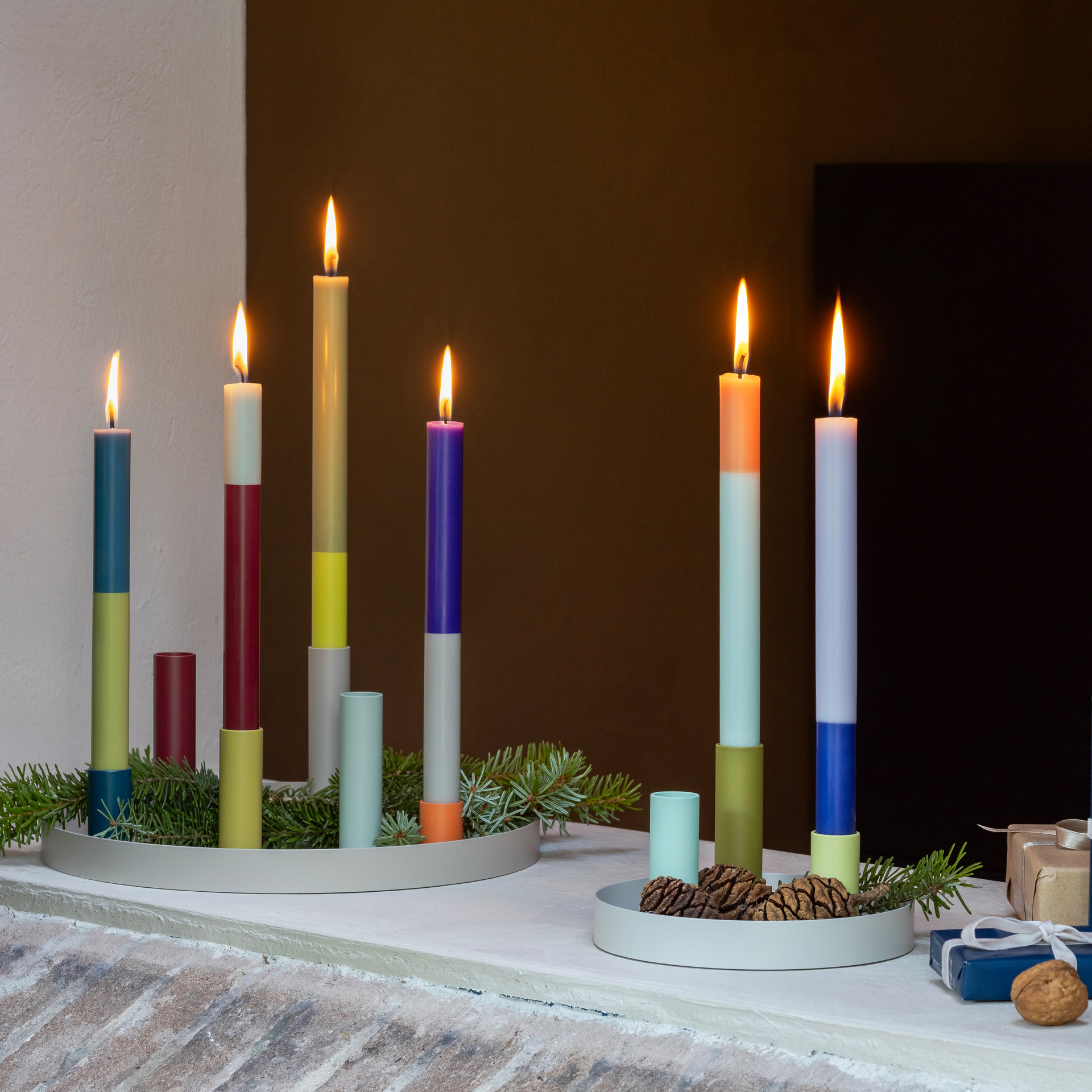 Brandende stokkaarsen 'Oslo' in een kleurrijk blokmotief, gerangschikt op ronde schalen met sparrentakken. Sommige kaarsen zijn verschillend van hoogte en sommige branden.