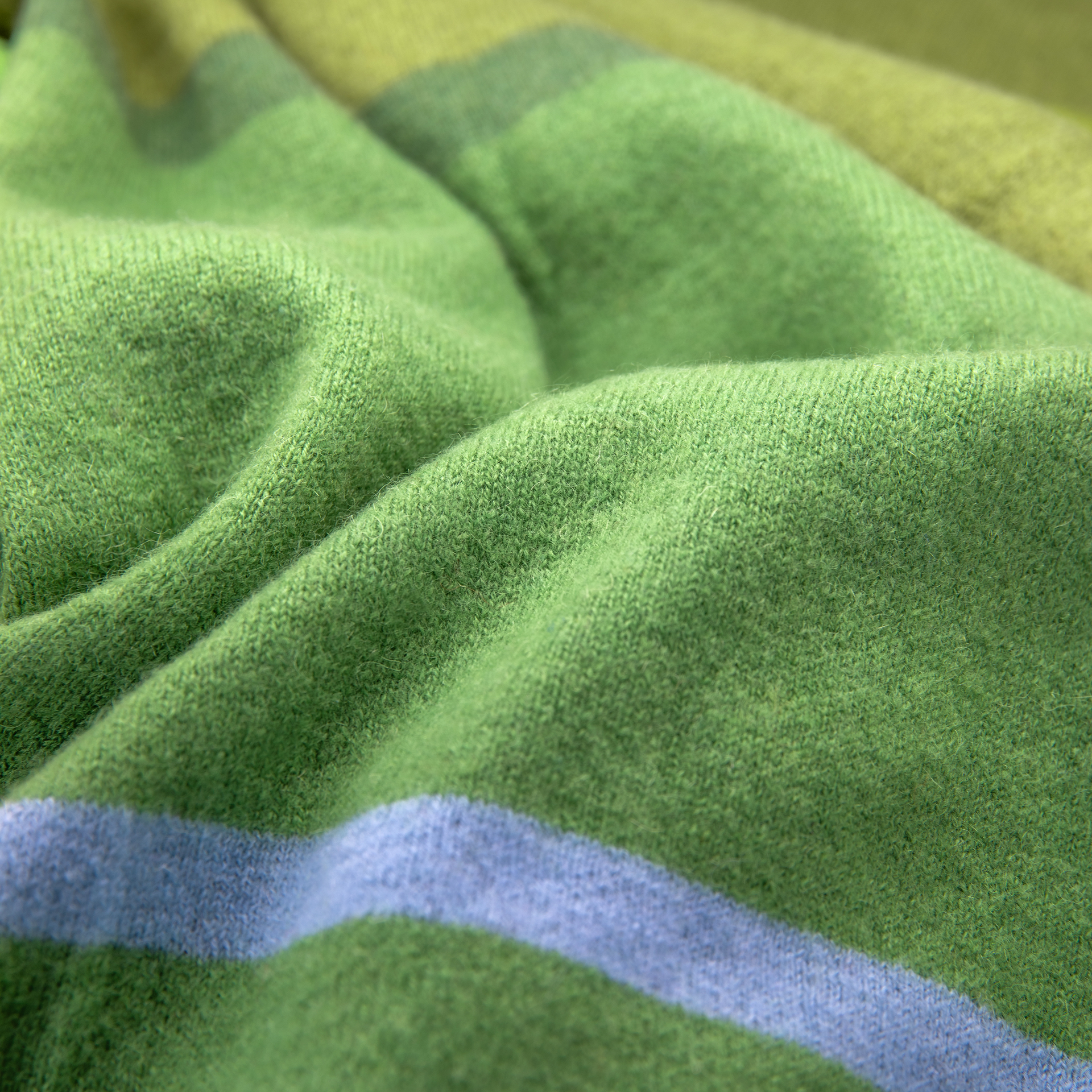 Primo piano di una texture morbida e intrecciata. Le strisce verdi e verde chiaro sono separate da una sottile linea blu. Il tessuto si presenta in morbide pieghe.