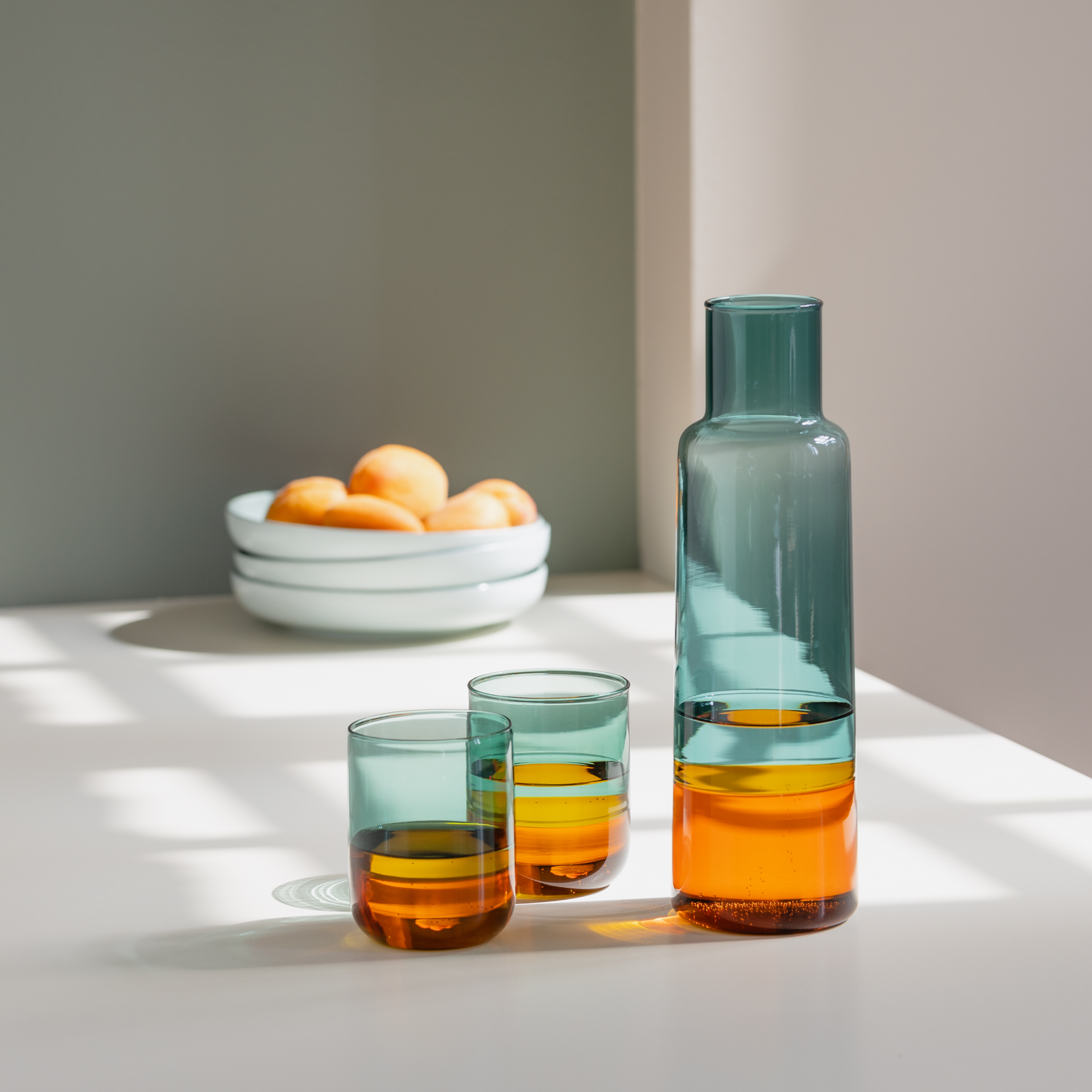 Two 'amber' glasses and a carafe stand on a white table. Filled with liquids in shades of amber and blue. A bowl of fruit in the background.