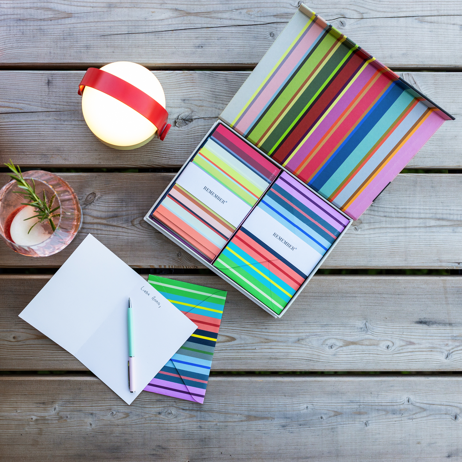 Ein offenes REMEMBER Kartenset mit bunten Klappkarten und gestreiften Umschlägen liegt in einer Magnetbox auf einem Holztisch. Daneben: geöffnete Karte mit "Liebe Anna,", Stift, kugelförmige Lampe mit rotem Band und Glas mit Rosmarindrink.