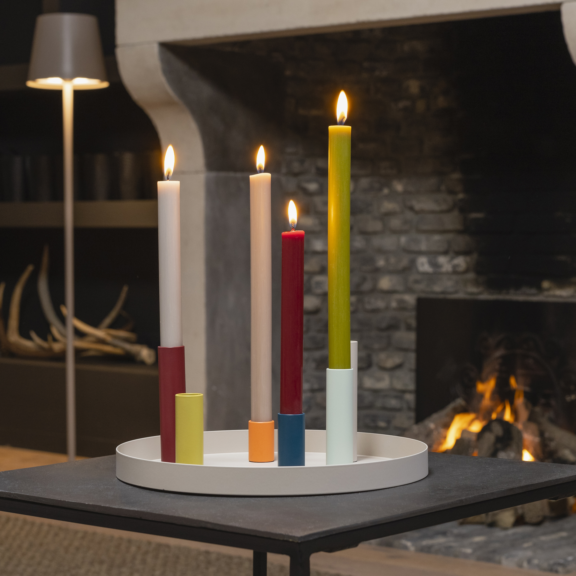 Il portacandele magnetico "Luna" si trova su un tavolo con candele accese di diversi colori. Sullo sfondo si vedono un camino e una lampada.