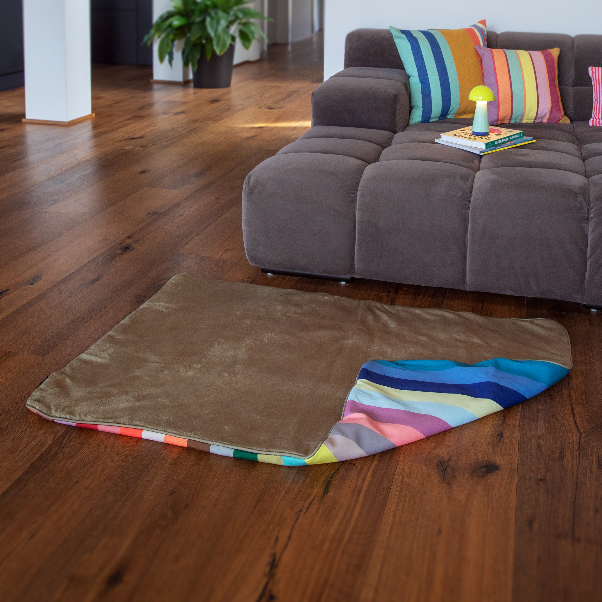 Eine braune, weiche Hundedecke liegt auf dunklem Holzboden im Wohnzimmer. Eine Ecke ist umgeschlagen und zeigt eine bunte Unterseite mit Streifen in Rosa, Blau, Gelb und Grün. Im Hintergrund steht ein graues Sofa.