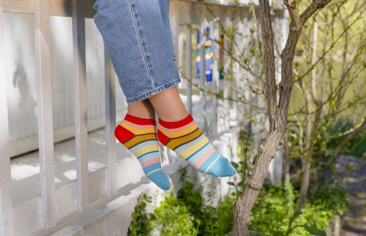 Eine Person trägt hellblaue Jeans und bunte Ringelsocken in Rot, Gelb, Braun, Hellblau, Rosa und Türkis. Die Füße baumeln entspannt über einem weißen Holzgeländer. Im Hintergrund: grüne Pflanzen und ein Baum in der Sonne.
