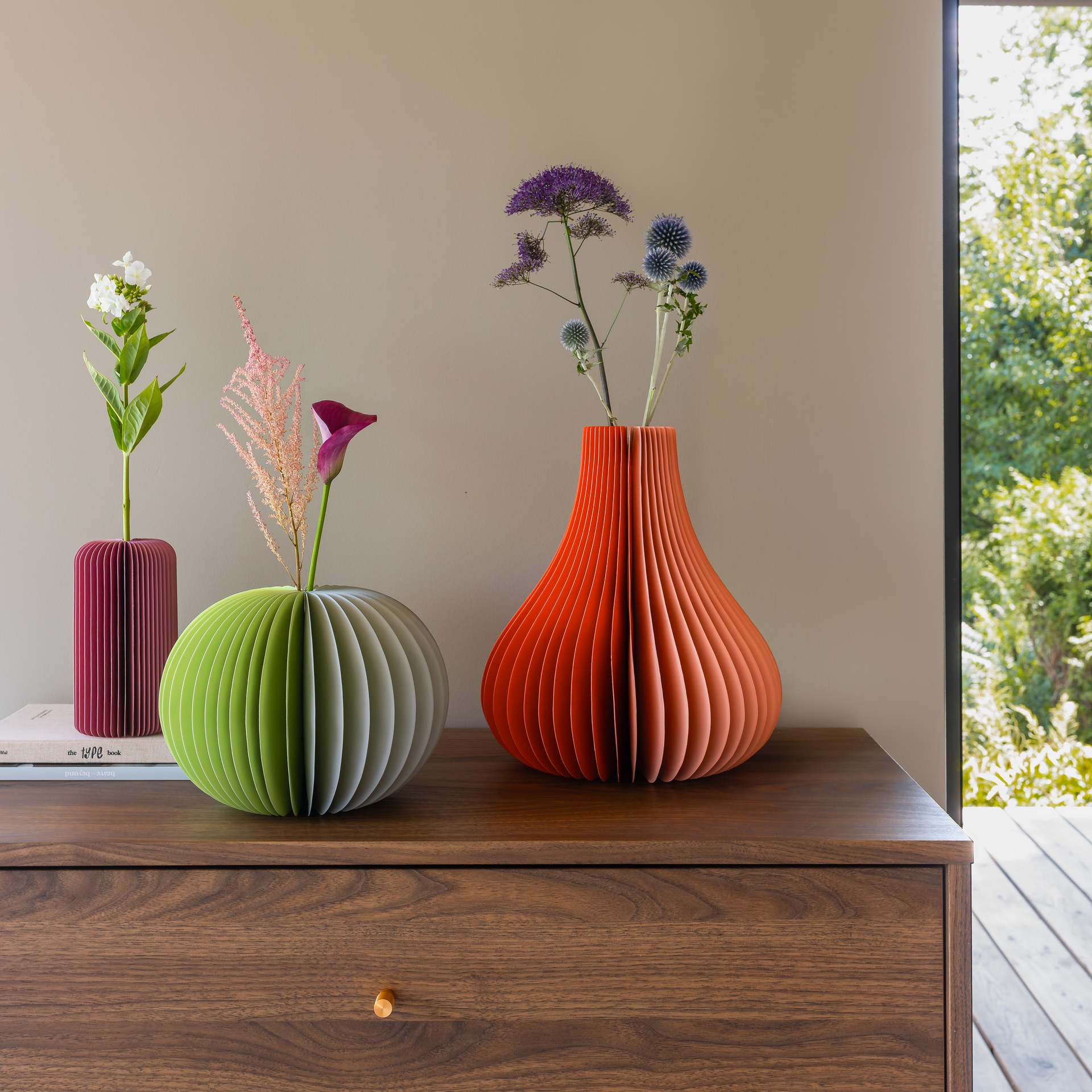 Trois vases en papier pliés sur une armoire sombre : à gauche, vase bordeaux à fleurs blanches ; au centre, vase vert clair avec calla rose ; à droite, grand vase orange « Yuki » aux chardons violets.