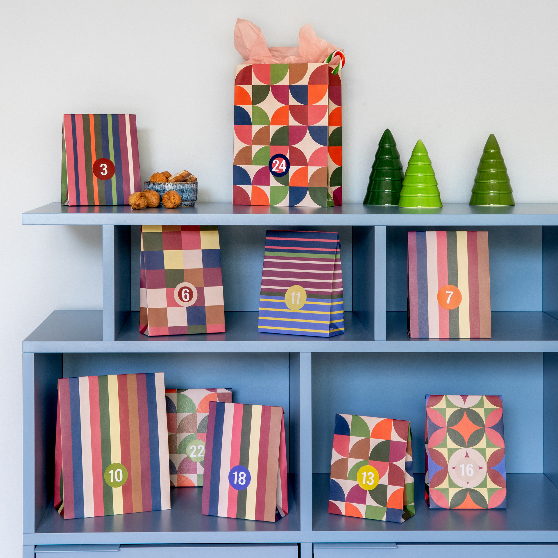 Bolsas de papel de colores para el calendario de Adviento con pegatinas con números del 1 al 24 sobre una estantería azul. Varios diseños: rayas, cuadros y motivos geométricos. Arriba: bolsa n.º 24, cuenco con frutos secos y tres árboles verdes.