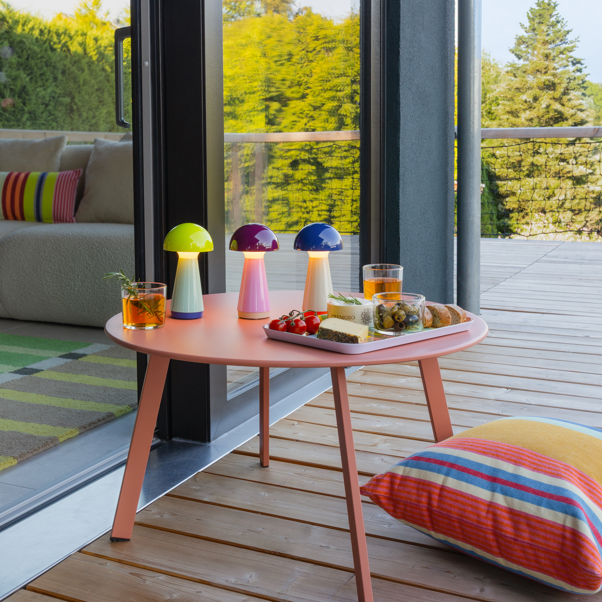 Trois lampes de table lumineuses (citron vert, baie, bleu) posées sur une table ronde couleur saumon, sur une terrasse en bois. À côté, des boissons et des en-cas. À l'arrière-plan, un canapé et des arbres derrière une porte vitrée.